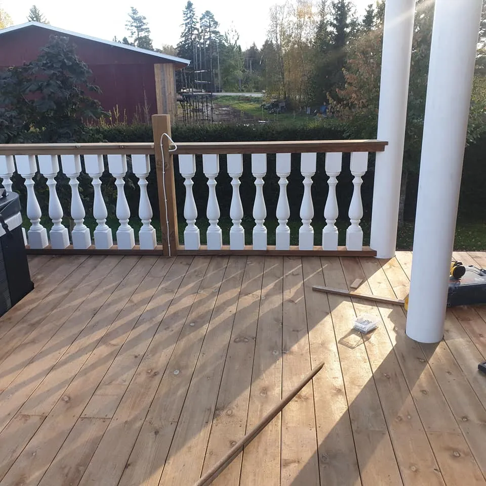 Aurinkoinen puuterassi, jossa on valkoinen kaide ja kaksi valkoista pylvästä. Pitkät varjot lankeavat terassille. Taustalla näkyy punainen rakennus ja puita.