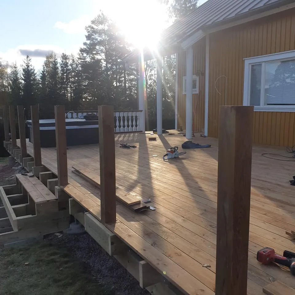 Aurinkoinen kuva keltaisesta puutalosta ja sen rakenteilla olevasta puisesta terassista. Terassilla on tumma poreallas, pystyssä olevia puupylväitä ja työkaluja. Vasemmalla rakennetaan portaita. Kirkas auringonvalo luo pitkiä varjoja terassin pinnalle. Taustalla on metsää.