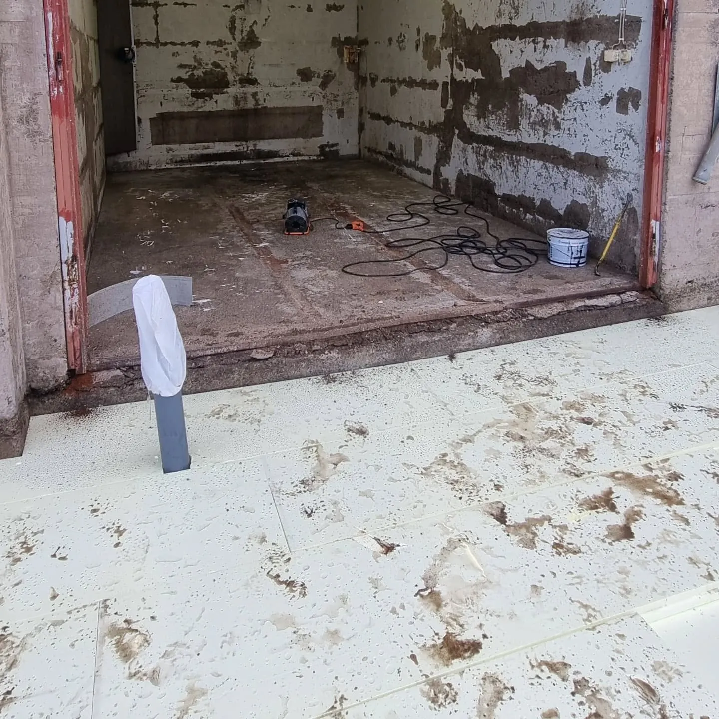 Kuva esittää avoimen, remontoidun tai purkutilassa olevan huoneen, jossa on osittain maalattuja betoniseiniä ja likainen betonilattia. Lattialla on musta-oranssi kone, sähköjohtoja, valkoinen maalipurkki ja keltainen työkalu. Huoneen edustalla on valkoisia levyjä, joissa on vesipisaroita ja tummanruskeita likaroiskeita. Vasemmalla etualalla harmaasta putkesta nousee valkoinen suojus.