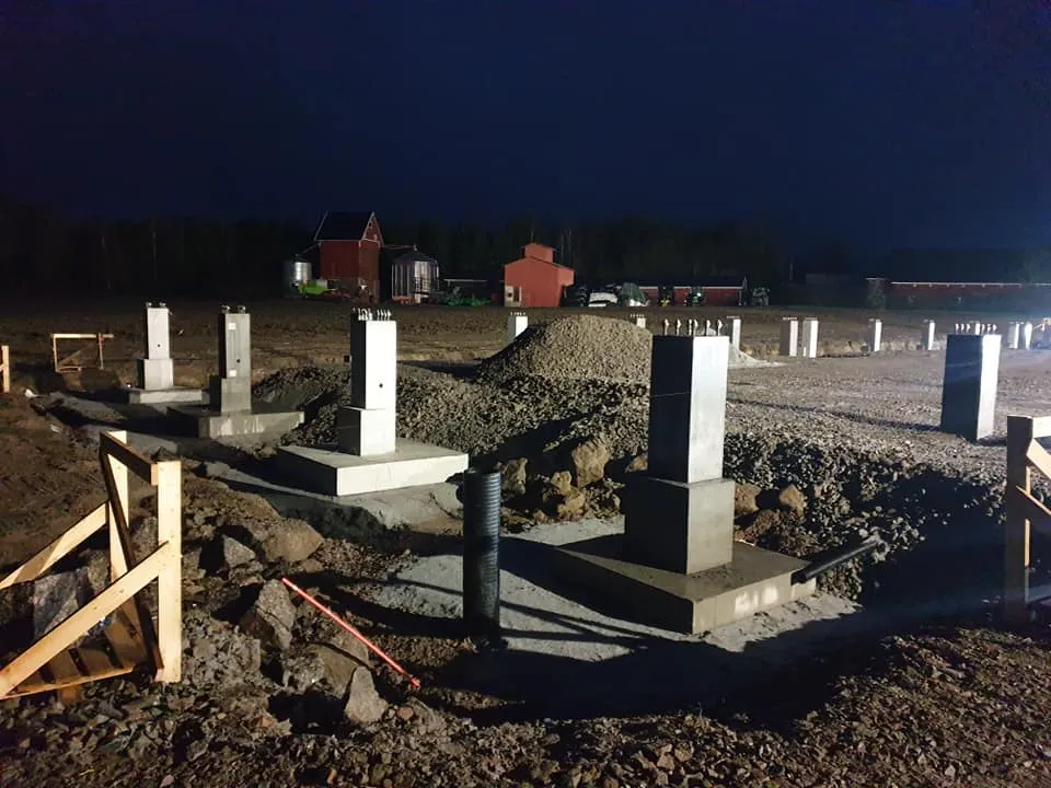 Yökuva rakennustyömaasta, jossa useita harmaita betonipilareita ja niiden anturoita on valmiina. Maassa on soraa ja kiviä, ja etualalla näkyy puurakenteita. Taustalla tumman taivaan alla on punaisia ja ruskeita rakennuksia sekä puita. Keinotekoinen valaistus luo teräviä varjoja.