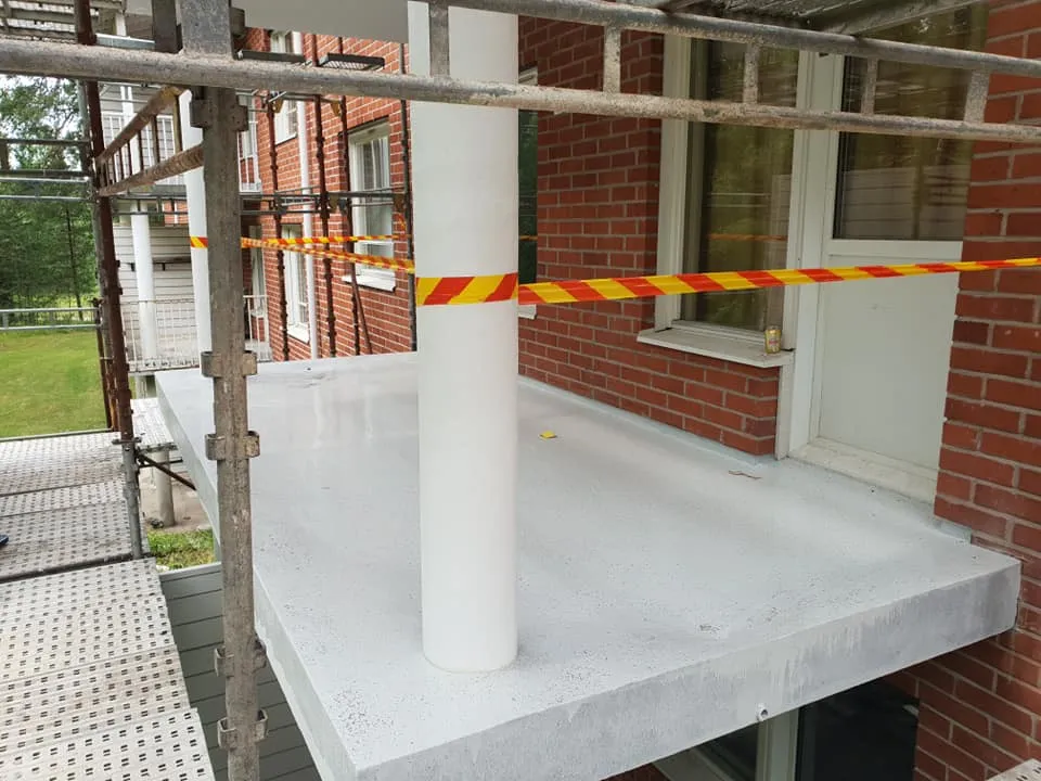 Vaaleanharmaa, juuri maalattu betoniterassi rakennustelineiden ja tiilirakennuksen edessä. Terassin poikki kulkee keltainen ja punainen raidallinen varoitusnauha. Terassilla on valkoinen pylväs ja ikkunalaudalla juomatölkki. Taustalla näkyy vihreää luontoa.