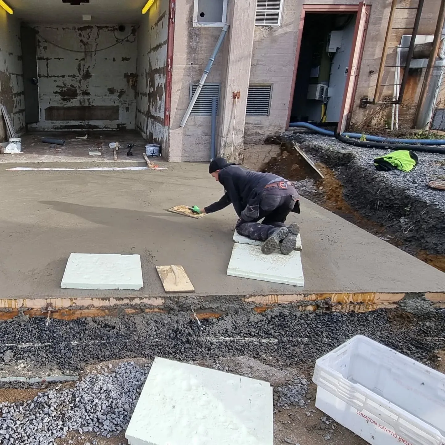 Mies polvillaan tasoittaa harmaata betonia hiertolastalla ulkona rakennuksen edessä. Hänellä on tummat työvaatteet ja musta pipo. Betonilaatan vieressä on soralla täytetty kaivanto. Taustalla näkyy vaaleanruskea rakennus avoimella oviaukolla, jonka sisällä on kuluneet seinät. Maassa on valkoisia eristelevyjä, työkaluja ja kirkkaanvihreä liivi.