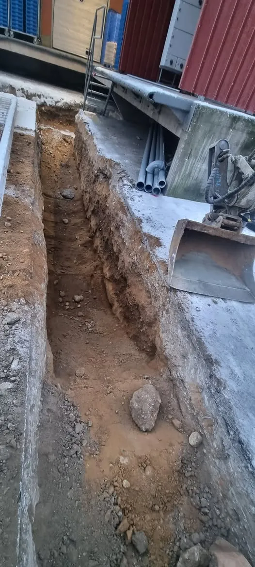 Pystysuuntainen kuva pitkästä, kapeasta kaivannosta, joka on kaivettu maahan. Kaivannon molemmilla puolilla on betonipäällysteiset reunat. Oikealla reunalla lepää kaivinkoneen metallinen kauha. Kaivannon takana ja oikealla ylhäällä näkyy rakennus, jossa on punaista aaltopeltiä ja sinisiä osia, sekä ramppi, jonka alla on harmaita putkia. Kaivannon pohjalla on ruskeaa maata ja erikokoisia kiviä. Kuva on otettu päivänvalossa.