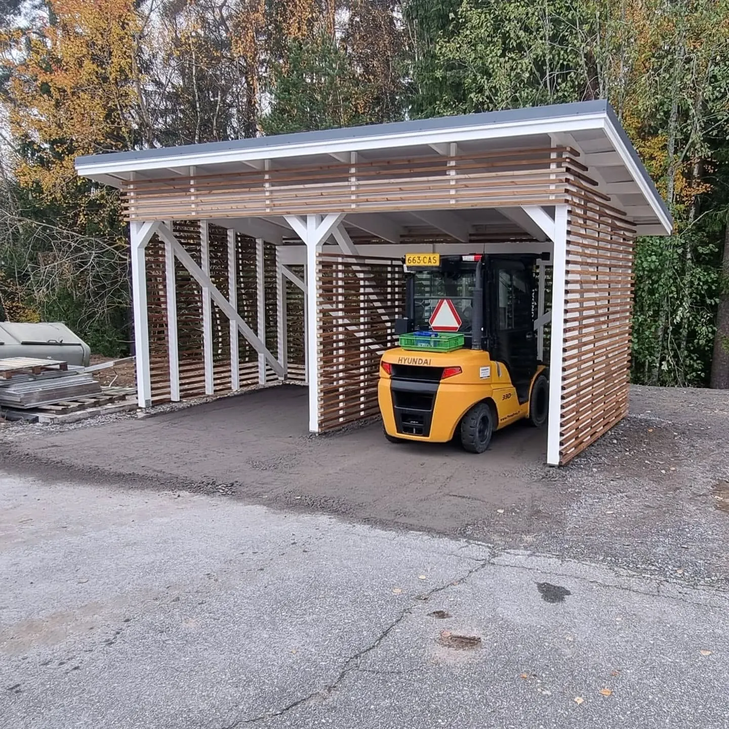 Uusi puinen katos, jossa on valkoiset tukipilarit ja vaaleanruskeat vaakasuorat puurimat seininä. Harmaakattoisen katoksen sisällä on pysäköity keltainen Hyundai-trukki, jonka lavalla on vihreä laatikko ja mastossa punainen varoituskolmio. Katoksen alla on tummaa soraa ja etualalla vaaleampaa asfalttia. Taustalla näkyy syksyisiä puita keltaisine ja vihreine lehtineen kirkkaassa päivänvalossa.