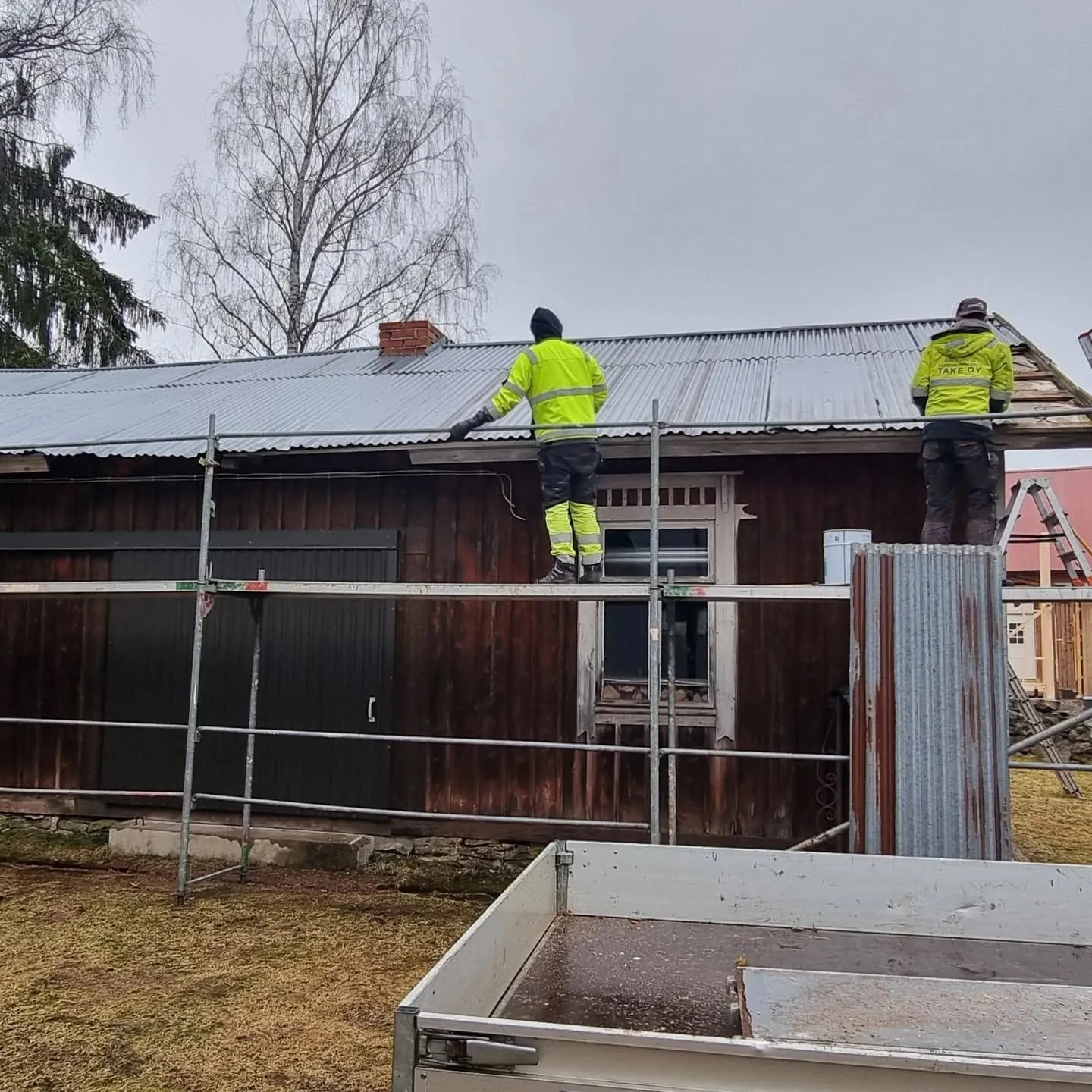 Kaksi työntekijää keltaisissa huomioliiveissä työskentelee tummanruskean puutalon aaltopeltikaton parissa. Toinen seisoo telineillä ja toinen tikkailla. Katolla on punainen tiilipiippu. Taustalla on puita ja harmaa pilvinen taivas. Etualalla näkyy valkoinen peräkärry.