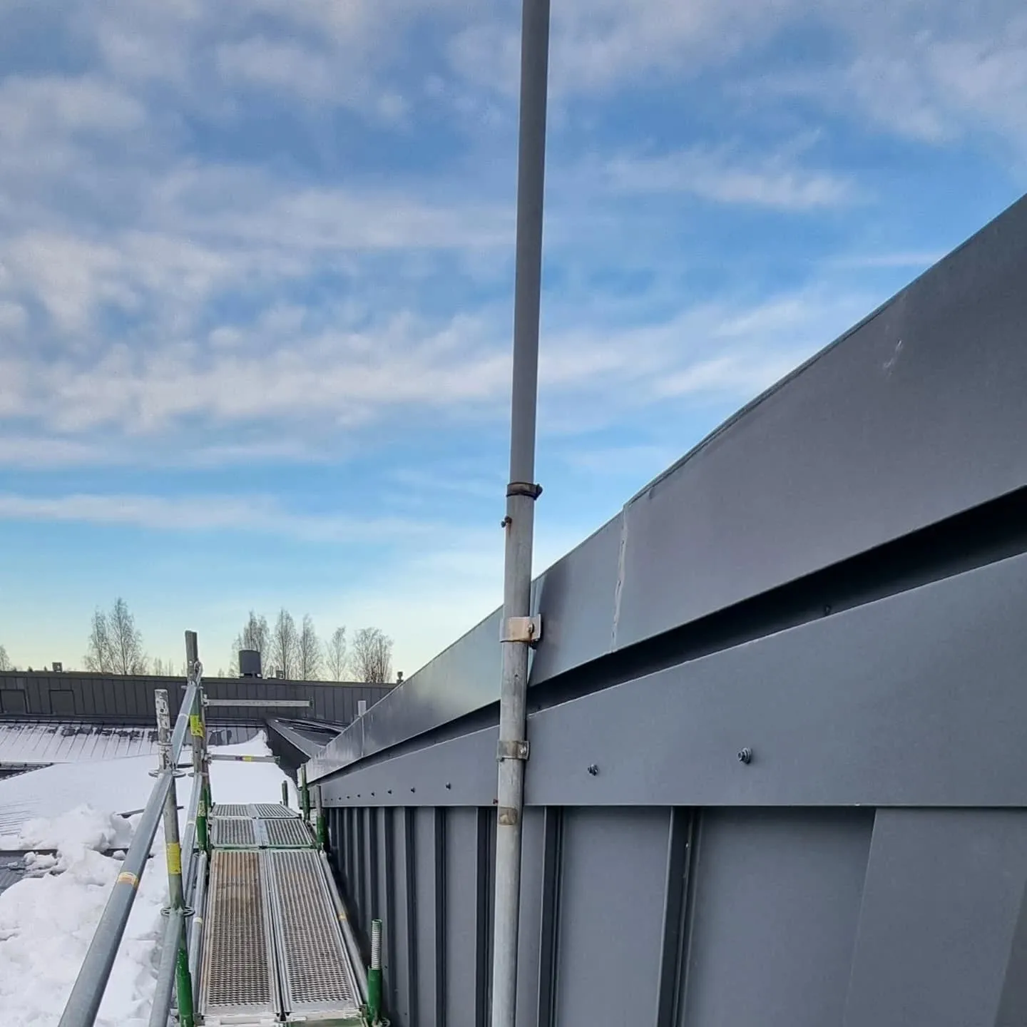 Kuva ylhäältäpäin rakennuksen tummanharmaasta metalliverhouksesta ja lumisesta katosta. Keskellä on pystysuora vaaleanharmaa pylväs kiinnitettynä seinään. Vasemmalla näkyy vihreä-harmaa rakennusteline. Taustalla on sininen taivas valkoisine pilvineen ja puita.