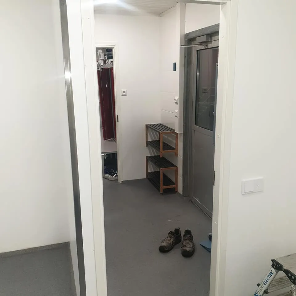 Käytävä, jossa on valkoiset seinät ja harmaa lattia. Vasemmalla on valkoinen seinä pystysuoralla metallinauhalla. Keskellä avoin ovi johtaa huoneeseen, jossa näkyy punaisia kaappeja. Oikealla on kaksi puista kenkätelinettä, hopeinen ovi lasipaneelilla ja valkoinen valokatkaisin. Lattialla on likaiset työkengät ja osa sinivalkoisesta työkalusta.