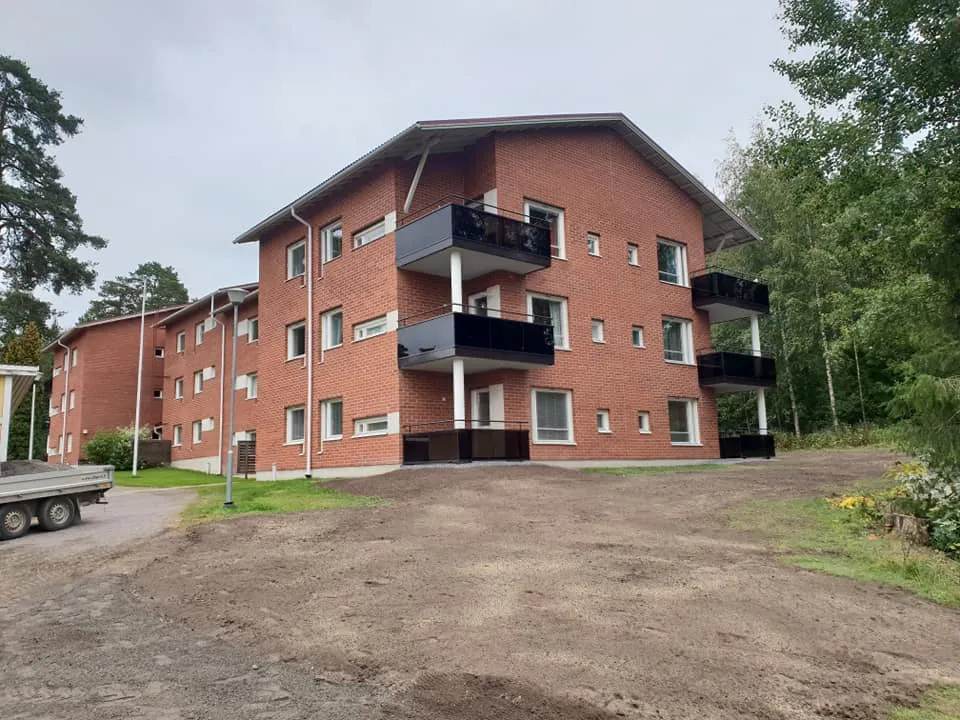 Punatiilinen asuintalo, jossa on mustat parvekkeet ja valkoiset ikkunat, seisoo hiekkaisen pihan edessä. Oikealla on tiheää metsää ja vasemmalla toinen samanlainen rakennus sekä peräkärryn osa. Taivas on pilvinen ja valo tasainen.