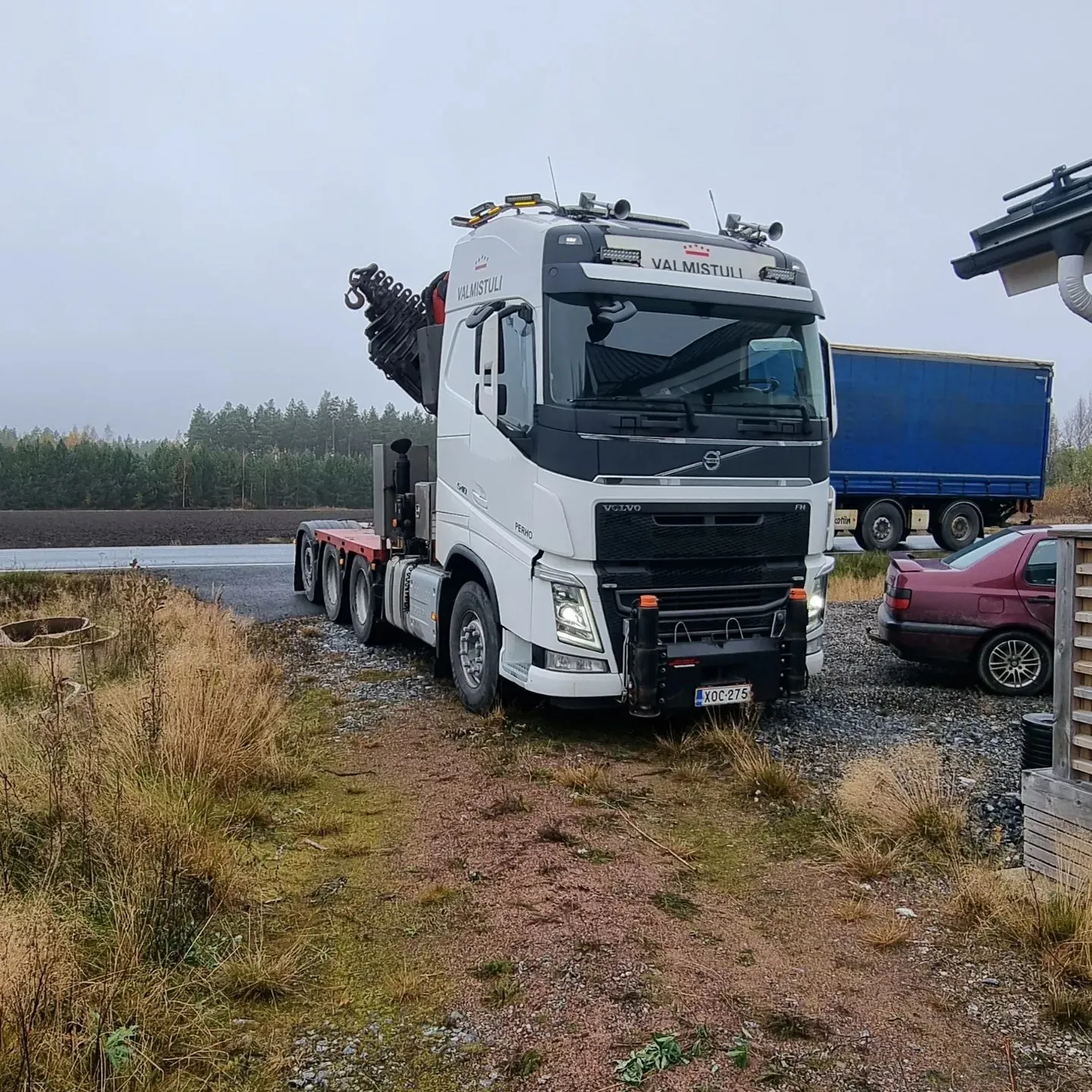 Valkoinen Volvo-kuorma-auto nosturilla on pysäköity soraiselle tielle pilvisenä päivänä. Kuorma-auton keulassa lukee 'VALMISTULI' ja rekisterikilpi on XOC-275. Taustalla näkyy metsää, pelto, sininen perävaunu ja punainen henkilöauto. Oikealla on osa puurakennusta ja maassa on kuivaa ruohoa.