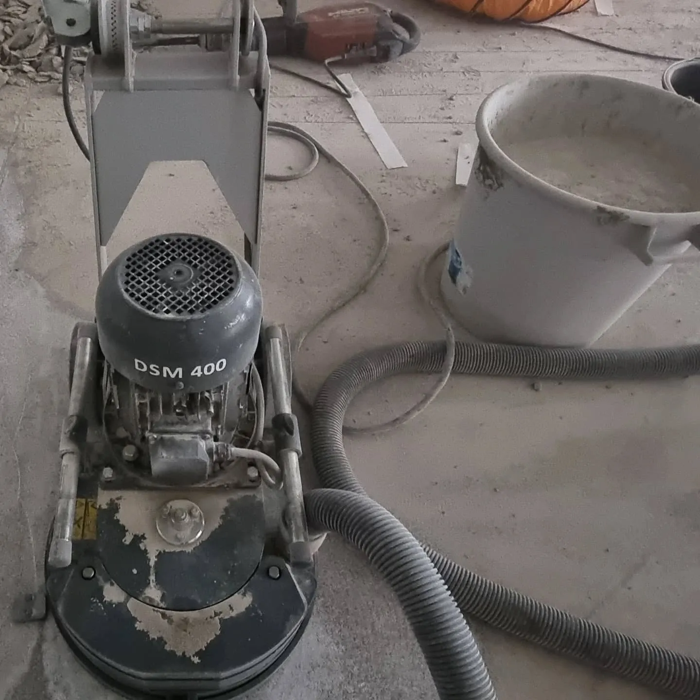 Pölyinen harmaa lattianhiontakone, jossa lukee "DSM 400", seisoo betonilattialla. Koneen vieressä on valkoinen ämpäri vaaleaa nestettä ja harmaa imuletku. Taustalla näkyy punainen työkalu ja oranssi putki.