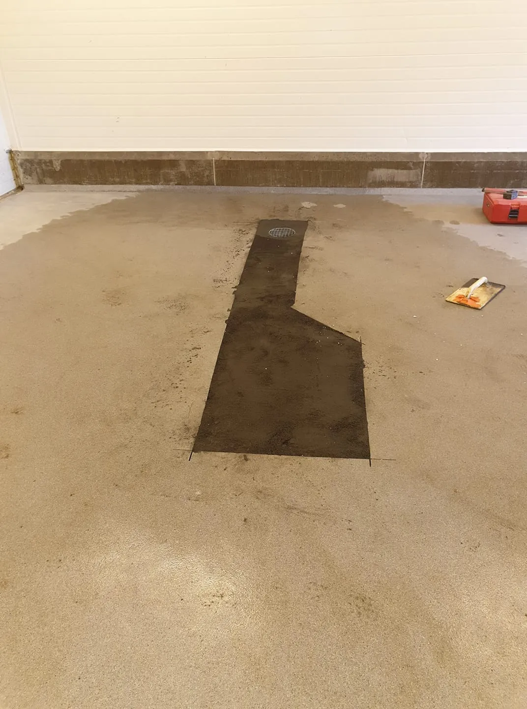 Vaalea betonilattia ja valkoinen paneeliseinä sisätilassa. Lattialla on L-muotoinen tumma laatta tai paikkaus, joka johtaa pyöreään metalliseen lattiakaivoon. Lattialla on kosteita alueita ja likaa. Oikealla on punainen työkalupakki ja likainen lasta.