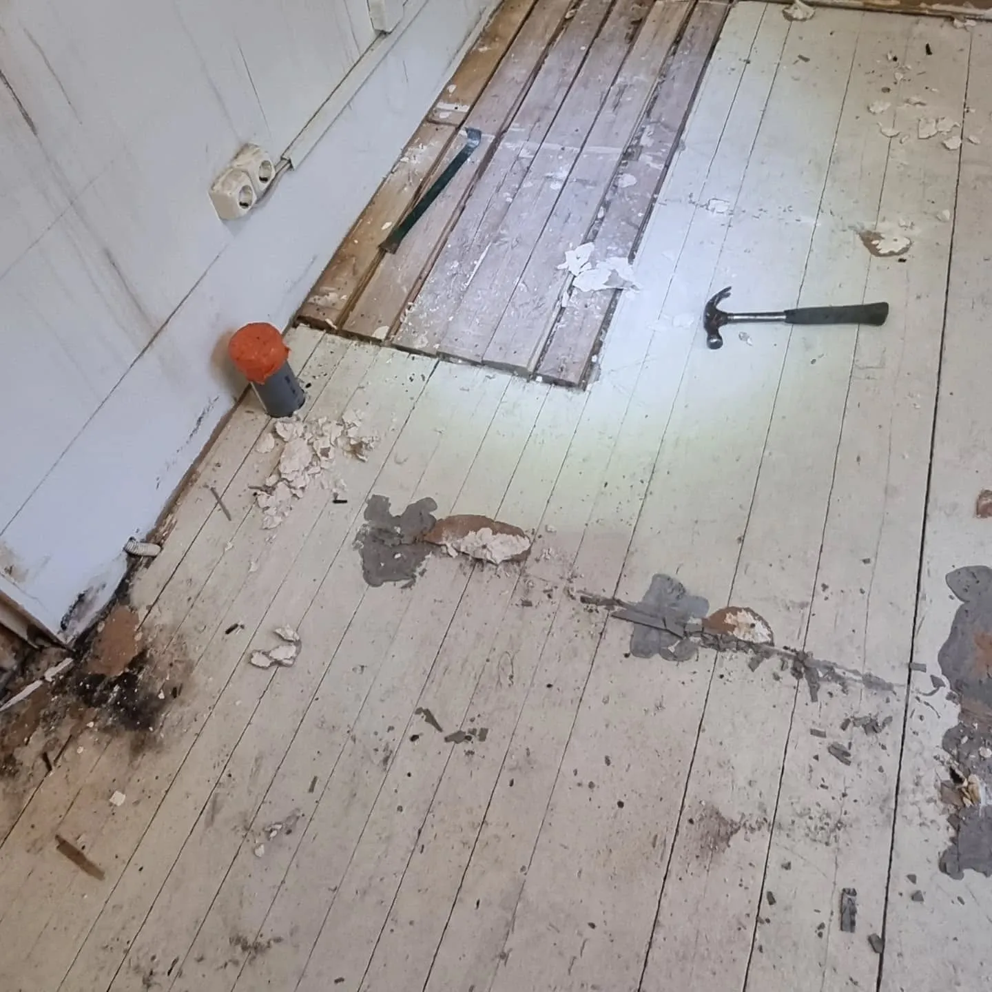 Kuva esittää remontin tai purkamisen alla olevaa huonetta. Lattialla on kuluneita, vaaleaksi maalattuja puulankkuja, joista osa on poistettu, paljastaen tummemman puun. Lattialla on likaa, roskia ja irronneita maalipalasia. Seinällä on kaksi vanhanaikaista pistorasiaa. Lattialla on harmaa putki oranssilla korkilla, sorkkarauta ja vasara.
