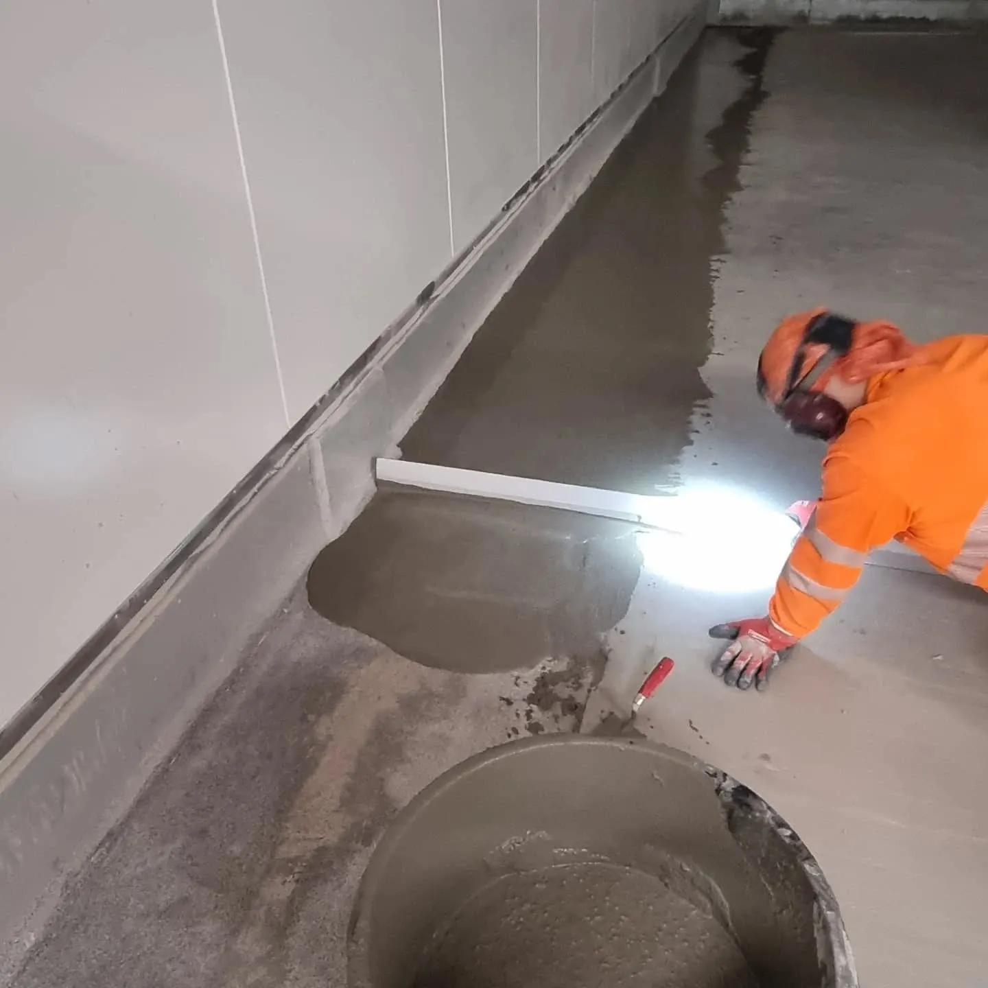 Työntekijä oransseissa työvaatteissa levittää harmaata lattiatasoitetta sisätiloissa. Hän on polvillaan ja käyttää kirkasta valoa työn apuna. Lattialla on valkoinen oikolauta ja punakahvainen lasta. Etualalla näkyy ämpäri täynnä tasoitetta. Seinät ovat vaaleanharmaat.