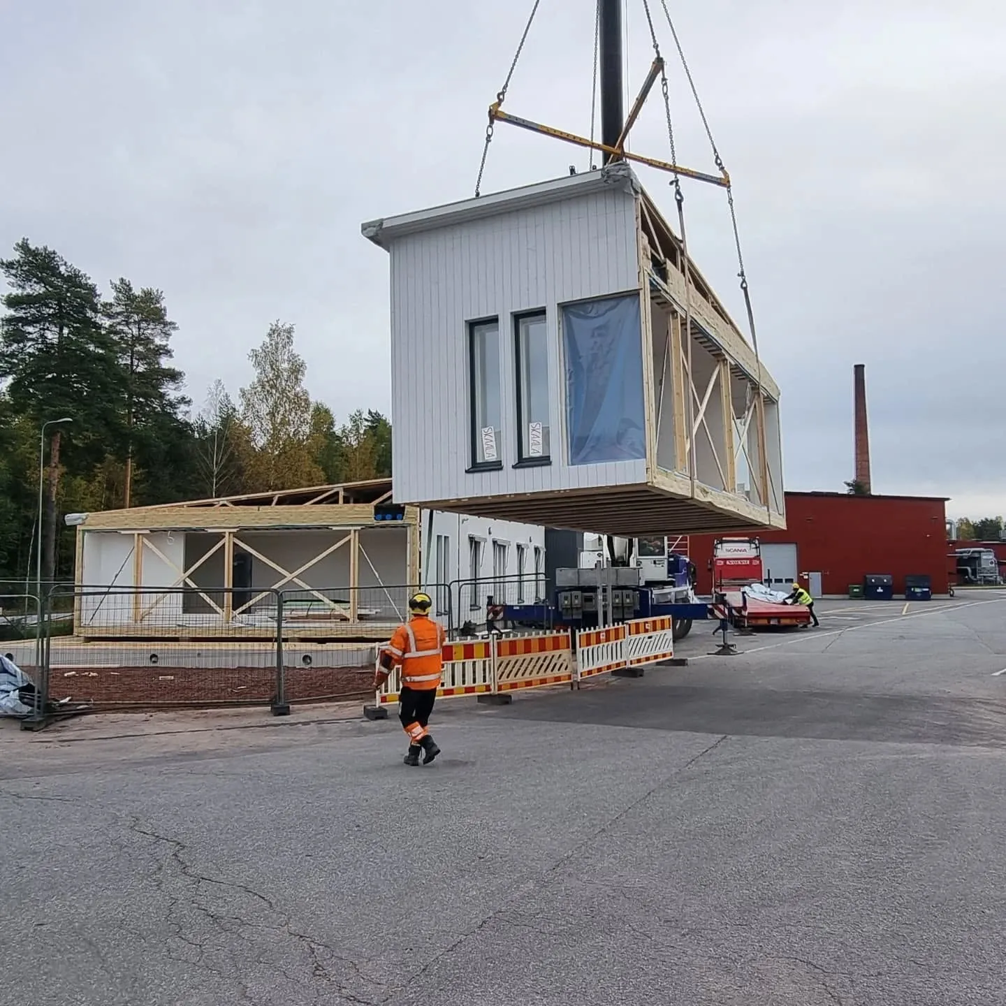 Nosturi nostaa valkoista esivalmistettua talomoduulia rakennustyömaalla. Moduulissa on mustakehyksiset ikkunat, joista yksi on peitetty sinisellä muovilla. Oranssiin huomioliiviin pukeutunut työntekijä kävelee asfaltilla etualalla. Taustalla näkyy keskeneräinen puurunkoinen rakennus, punainen tehdasrakennus ja korkea tiilisavupiippu pilvisen taivaan alla.