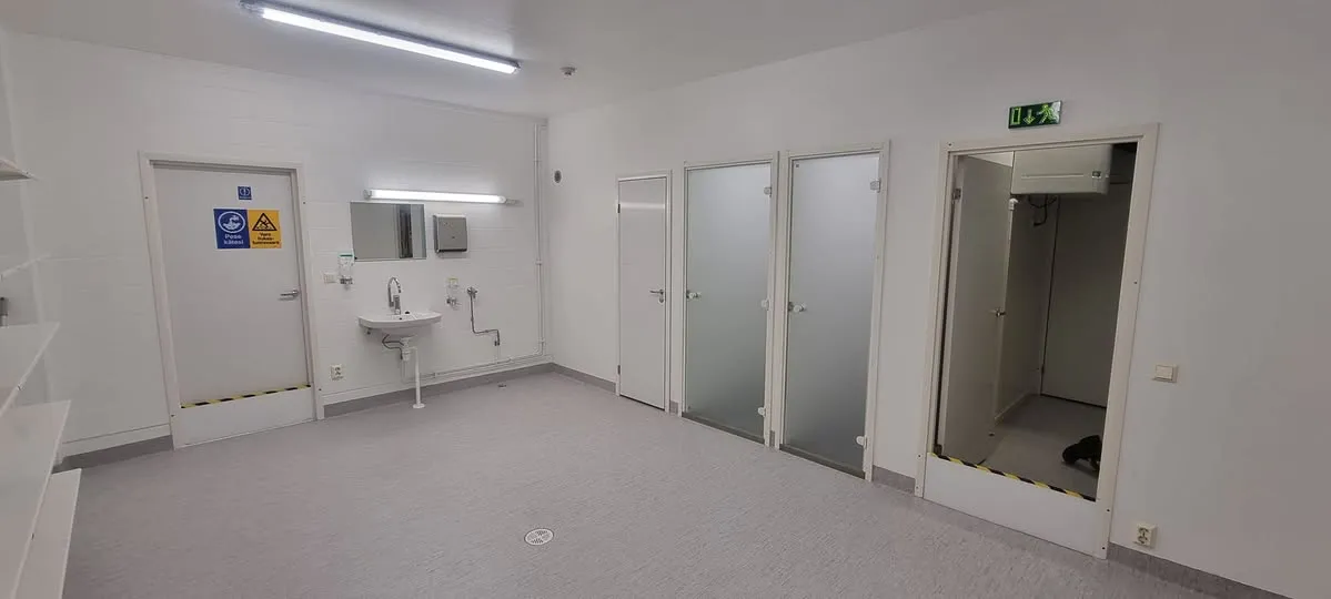 Avara ja valoisa huone, jossa on valkoiset seinät ja vaaleanharmaa lattia. Vasemmalla on valkoisia hyllyjä. Takaseinällä on valkoinen ovi, jossa on "Pese kätesi" ja "Varo liukasta lattiaa" -kyltit, sekä pesuallas peilillä ja saippua-annostelijalla. Oikealla on kolme ovea, joista kaksi on himmeälasisia, ja avoin oviaukko vihreällä poistumistiekyltillä.