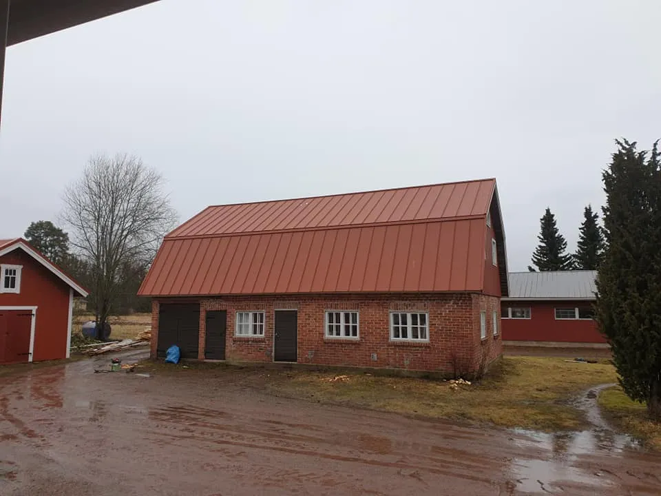 Keskellä kuvaa on punatiilinen rakennus punaisella peltikatolla, tummilla ovilla ja valkoisilla ikkunoilla. Vasemmalla on punainen puurakennus ja paljas puu. Oikealla näkyy havupuita ja taustalla toinen punainen rakennus harmaalla katolla. Märkä hiekkatie ja ruohoinen maa etualalla pilvisenä päivänä.