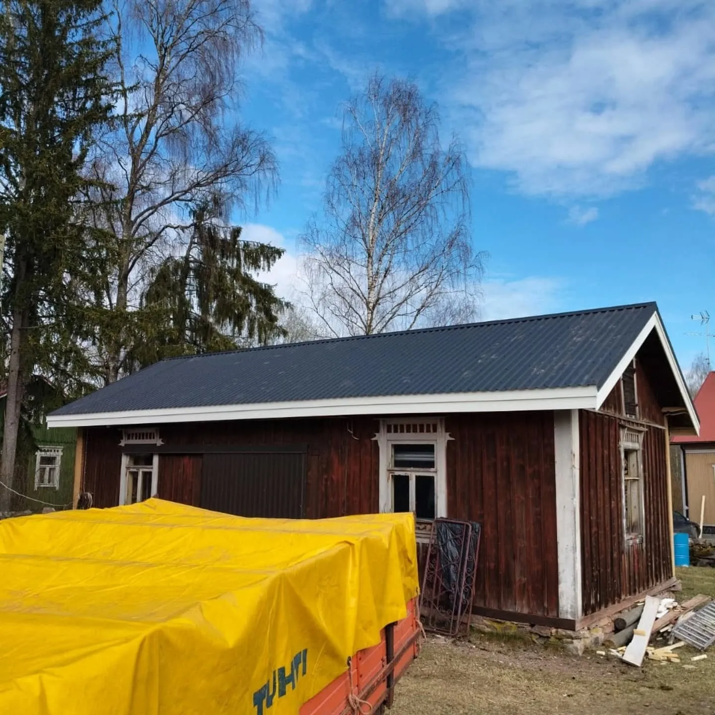 Aurinkoinen kuva vanhasta tummanpunaisesta puurakennuksesta, jossa on tummanharmaa aaltopeltikatto ja valkoiset räystäslaudat. Rakennuksen edessä on keltainen pressu peittämässä peräkärryä. Taustalla näkyy paljaita puita ja kirkkaan sininen taivas valkoisine pilvineen. Maassa on kuivaa ruohoa ja rakennusjätettä.