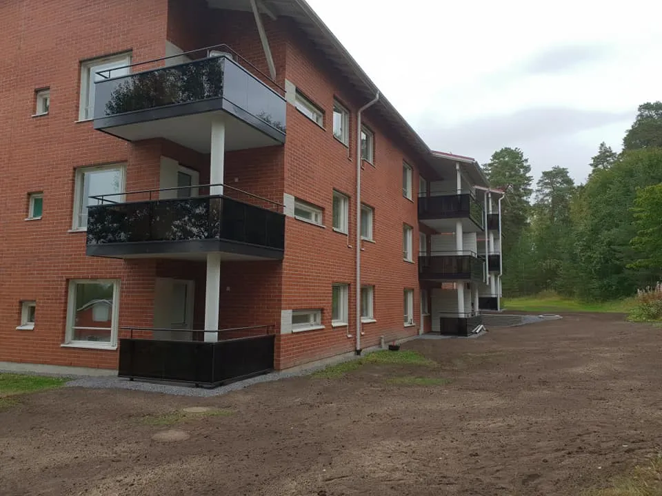 Punainen tiilirakennus, jossa on mustat parvekkeet ja valkoiset pilarit, seisoo ruskean maan ja vihreän metsän edessä pilvisenä päivänä.