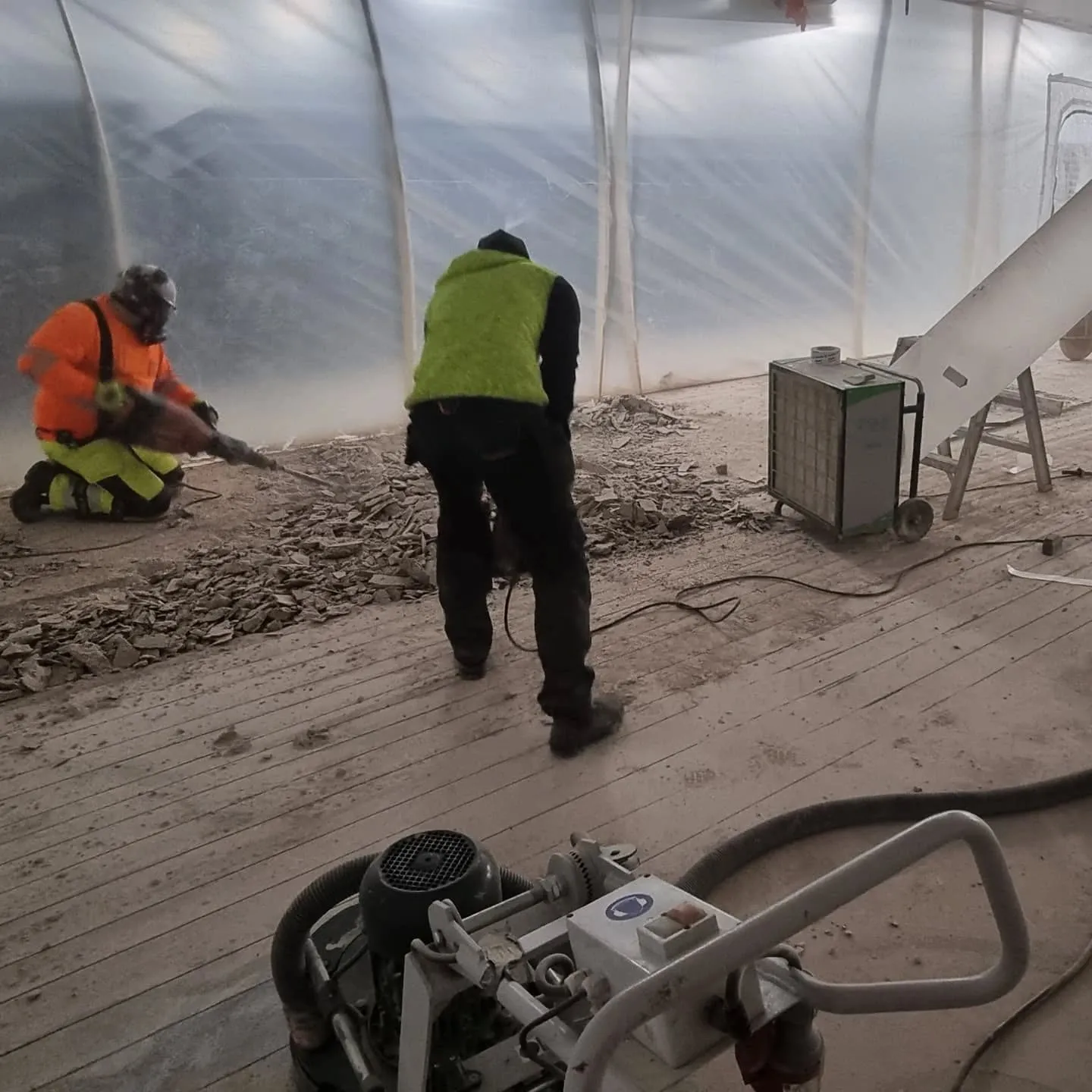 Kaksi työntekijää purkutyömaalla sisällä läpikuultavassa teltassa. Vasemmalla oranssipaitainen työntekijä polvillaan piikkaa lattiaa hengityssuojain kasvoillaan. Oikealla toinen työntekijä vihreässä liivissä seisoo selin. Lattialla on paljon purkujätettä ja pölyä. Keskellä näkyy pyörillä oleva pölynerotin ja etualalla lattianhiomakone.