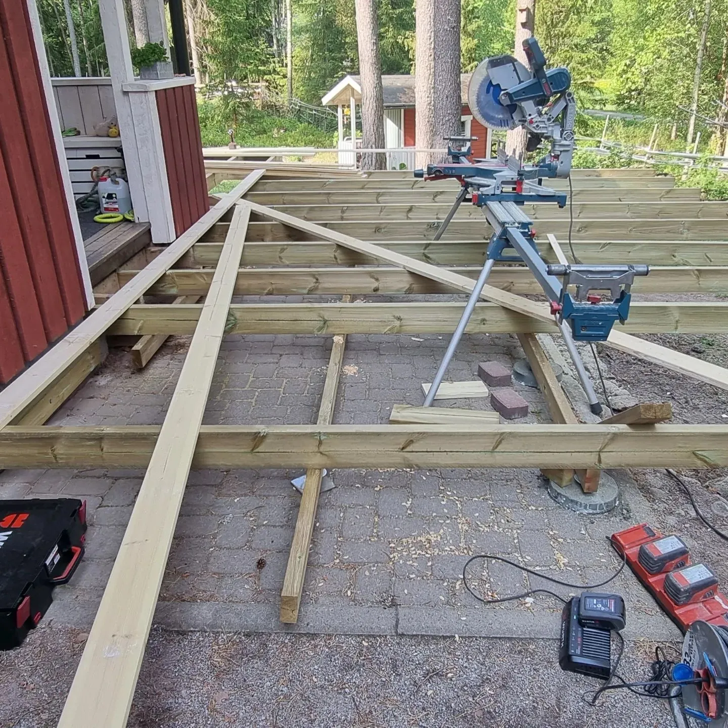 Ulkokuva terassin rakennustyömaasta päivänvalossa. Kuvan keskellä ja etualalla on vaaleanvihreästä puusta rakennettu terassin runko ja poikkipalkit. Oikealla on sininen ja hopeinen katkaisusaha omalla jalustallaan. Vasemmalla näkyy punainen talo valkoisilla reunuksilla. Maassa on harmaata kiveystä ja hiekkaa, sekä erilaisia työkaluja: musta työkalulaatikko, akkulaturi, punaisia akkuja ja jatkojohtokela. Taustalla on vihreää metsää ja pieni valkoinen rakennus.