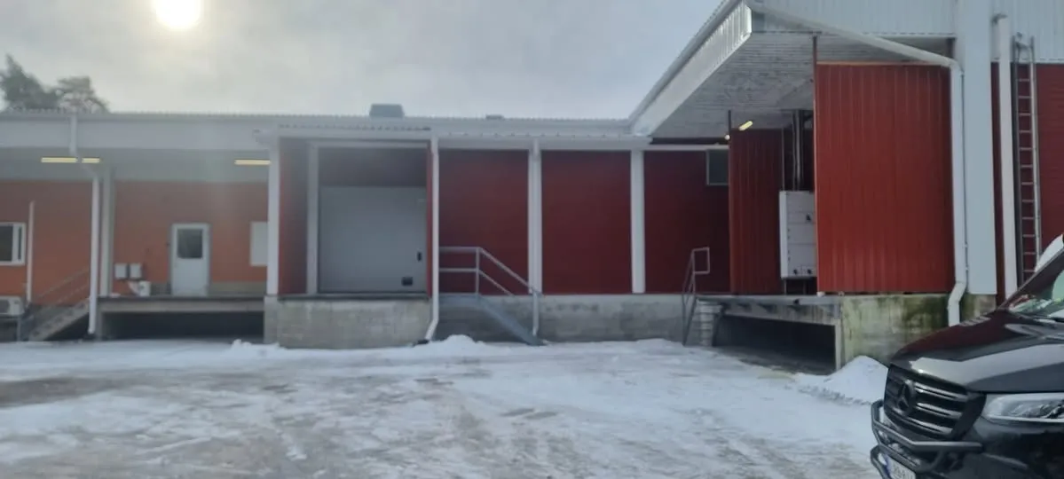Talvinen kuva varastorakennuksesta, jossa on oransseja ja punaisia seinäpintoja sekä valkoisia pilareita ja katosrakenteita. Maassa on lunta ja jäätä. Rakennuksessa on useita lastauslaitureita; yksi valkoinen nosto-ovi on auki. Oikealla etualalla näkyy mustan pakettiauton keula. Taivaalla on kirkas, sumuinen aurinko.