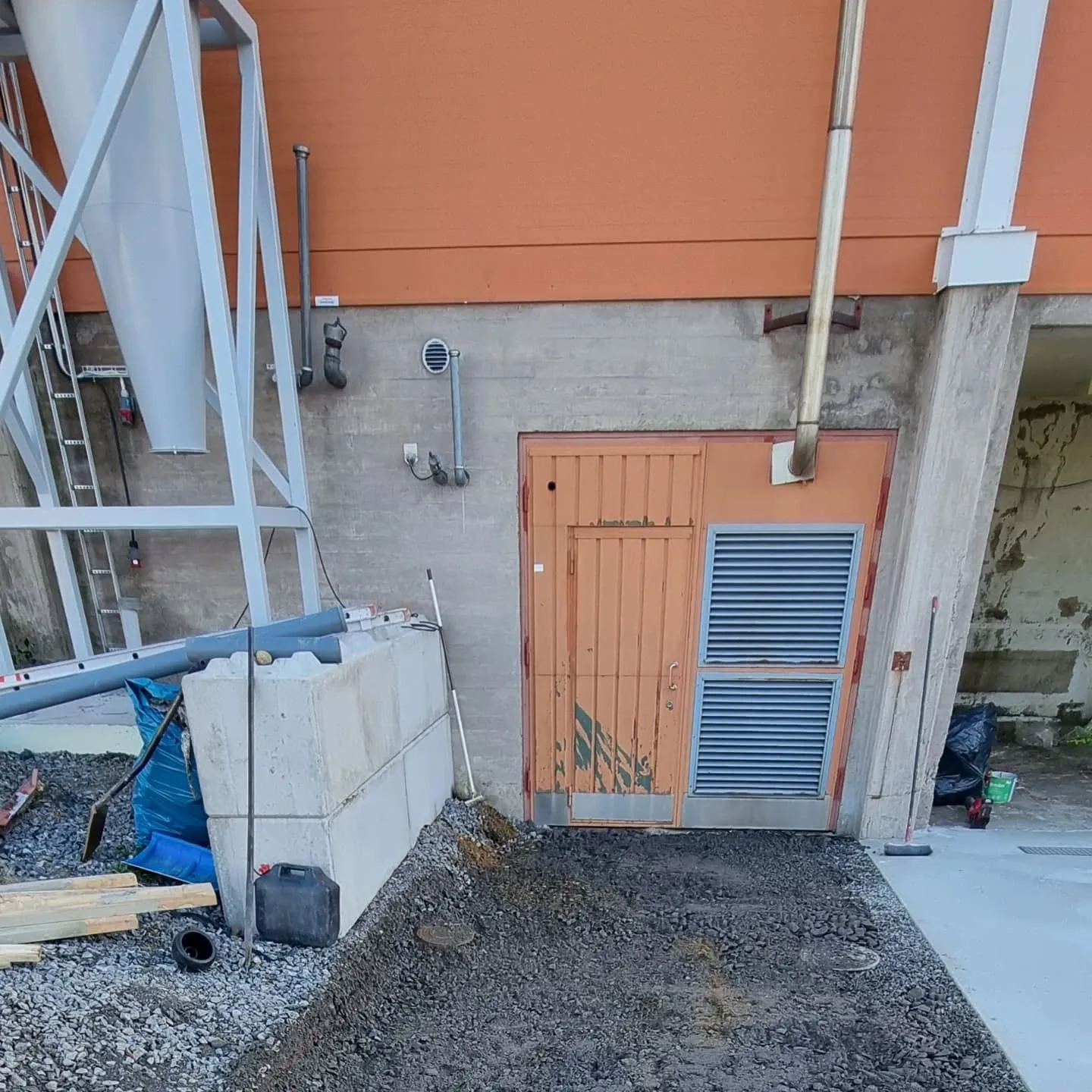 Kuva esittää rakennuksen ulkoseinää, jossa on oranssi yläosa ja harmaa betoninen alaosa. Vasemmalla on valkoinen kartiomainen siilo harmaassa metallirungossa. Betoniseinässä on oranssi metalliovi, jonka alla on kaksi sinistä ilmanvaihtoritilää. Oven yläpuolella ja seinällä on erilaisia putkia ja tuuletusaukkoja. Maassa oven edessä on tummaa soraa, ja oikealla on vaalea betonikäytävä. Vasemmalla maassa on rakennustarvikkeita, kuten suuria betonilohkoja, putkia, sininen pressu ja puutavaraa.