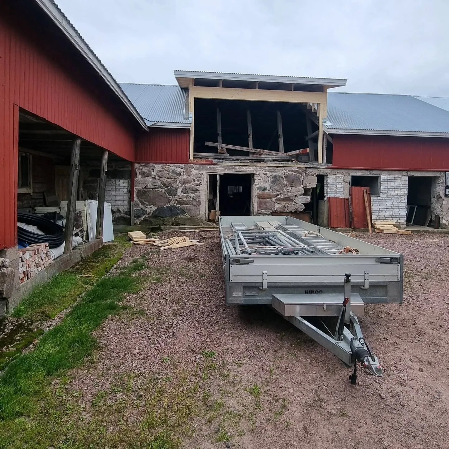 Vanha punainen rakennus, jossa on kivijalka ja aaltopeltikatto, on remontissa. Katolla on uusi puurakennelma. Etualalla on hopeinen peräkärry täynnä rakennustelineitä soraisella pihalla pilvisenä päivänä.