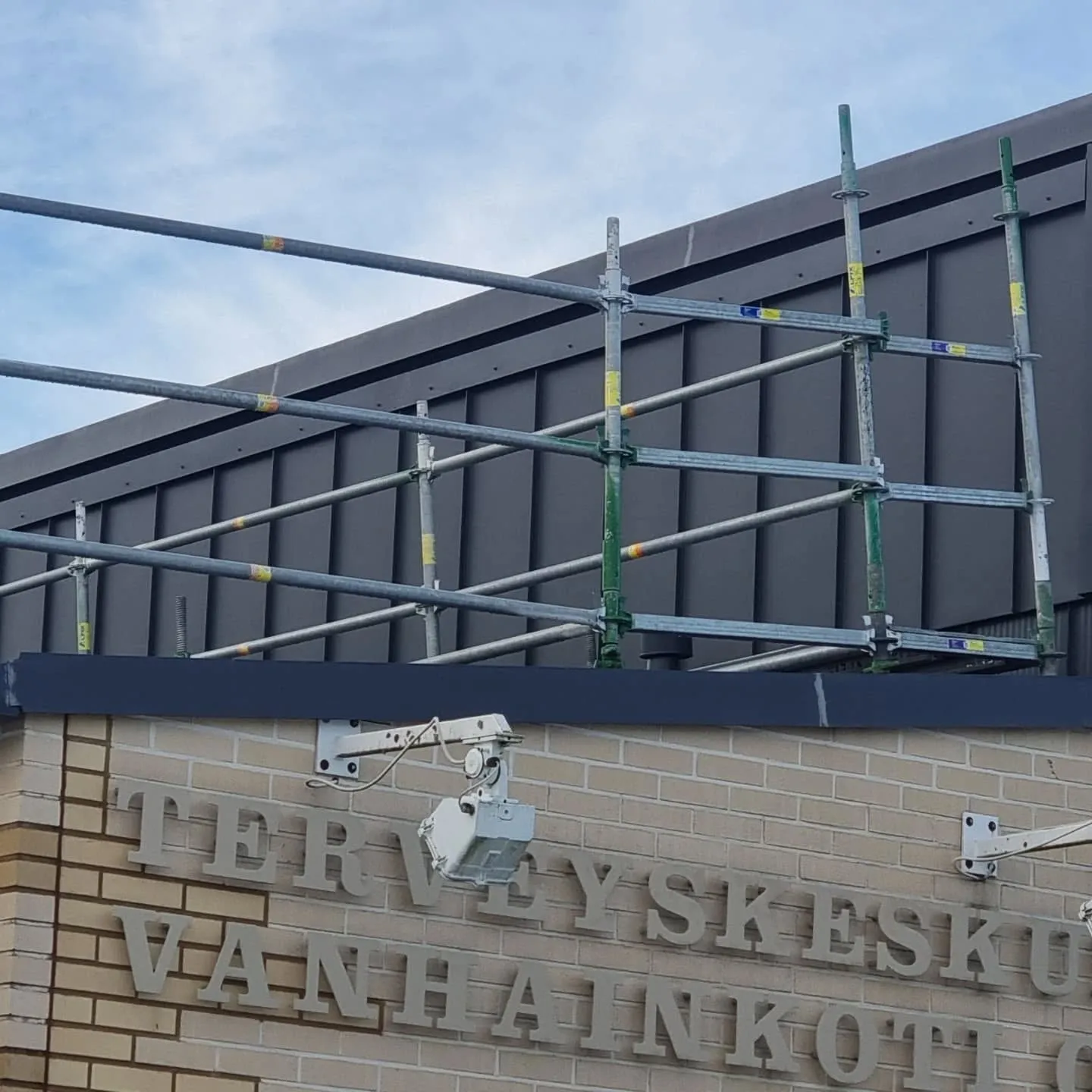 Kuva rakennuksen yläosasta, jossa on vaaleanruskea tiiliseinä ja tummanharmaa pystypaneelinen julkisivu. Tiiliseinällä on vaaleita kirjaimia, jotka muodostavat osan tekstistä "TERVEYSKESKUS VANHAINKOTI". Kirjainten edessä on valkoinen, varren päässä oleva laite. Rakennuksen edessä on metalliset rakennustelineet, joissa on harmaita ja vihreitä putkia sekä keltaisia ja sinisiä tarroja. Taustalla on sininen pilvinen taivas.