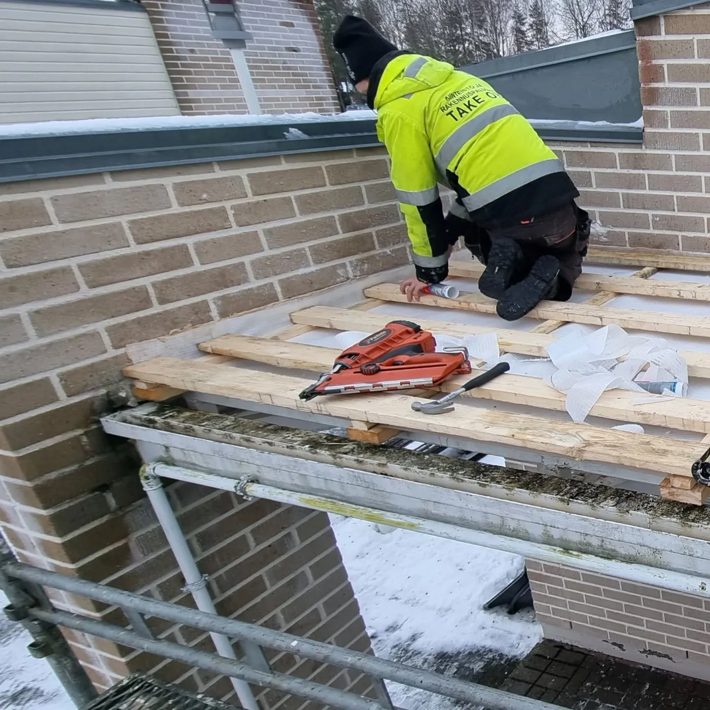 Työntekijä keltaisessa ja mustassa työtakissa on polvillaan puurakenteen päällä ulkona lumisessa ympäristössä. Hän levittää ainetta tuubista. Lähistöllä on naulapyssy ja vasara. Taustalla näkyy tiiliseinä ja vesikouru.