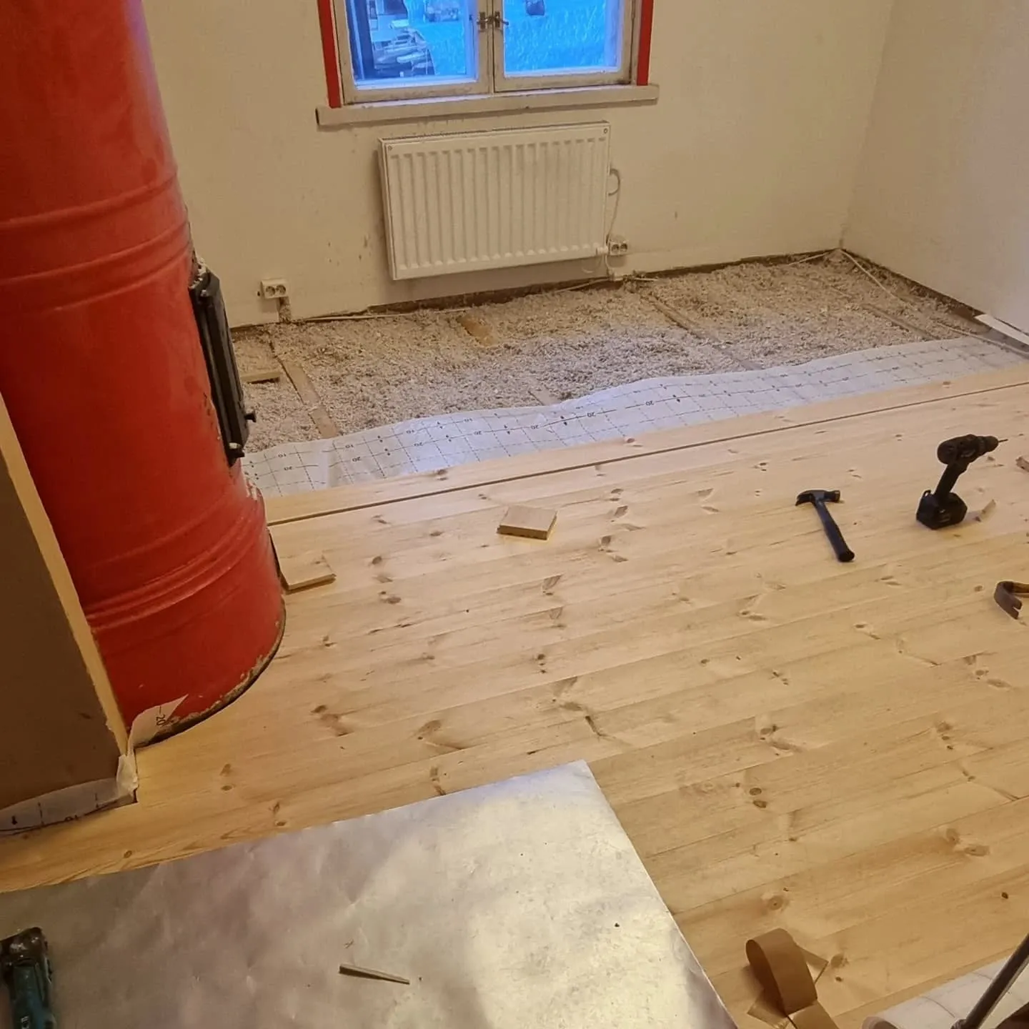 Huoneen lattian asennus on käynnissä. Etualalla on vaaleita puulattialautoja, ja taustalla näkyy vaaleaa eristemateriaalia ja ruudukollinen alusmateriaali. Vasemmalla on korkea, punainen pönttöuuni mustalla luukulla. Oikealla lattialla on vasara ja porakone. Taustalla on valkoinen seinä, punakehyksinen ikkuna ja valkoinen patteri. Huoneessa on kirkas sisävalaistus.