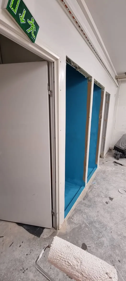 Valkoisessa huoneessa, jossa on harmaa betonilattia, näkyy vihreä poistumiskyltti valkoisen oven yläpuolella. Ovi on hieman raollaan. Oikealla seinässä on kolme syvennystä, joiden sisäpinnat on maalattu kirkkaan sinisiksi. Lattialla on valkoinen maalitela, ja oikealla taustalla näkyy polvistunut henkilö.