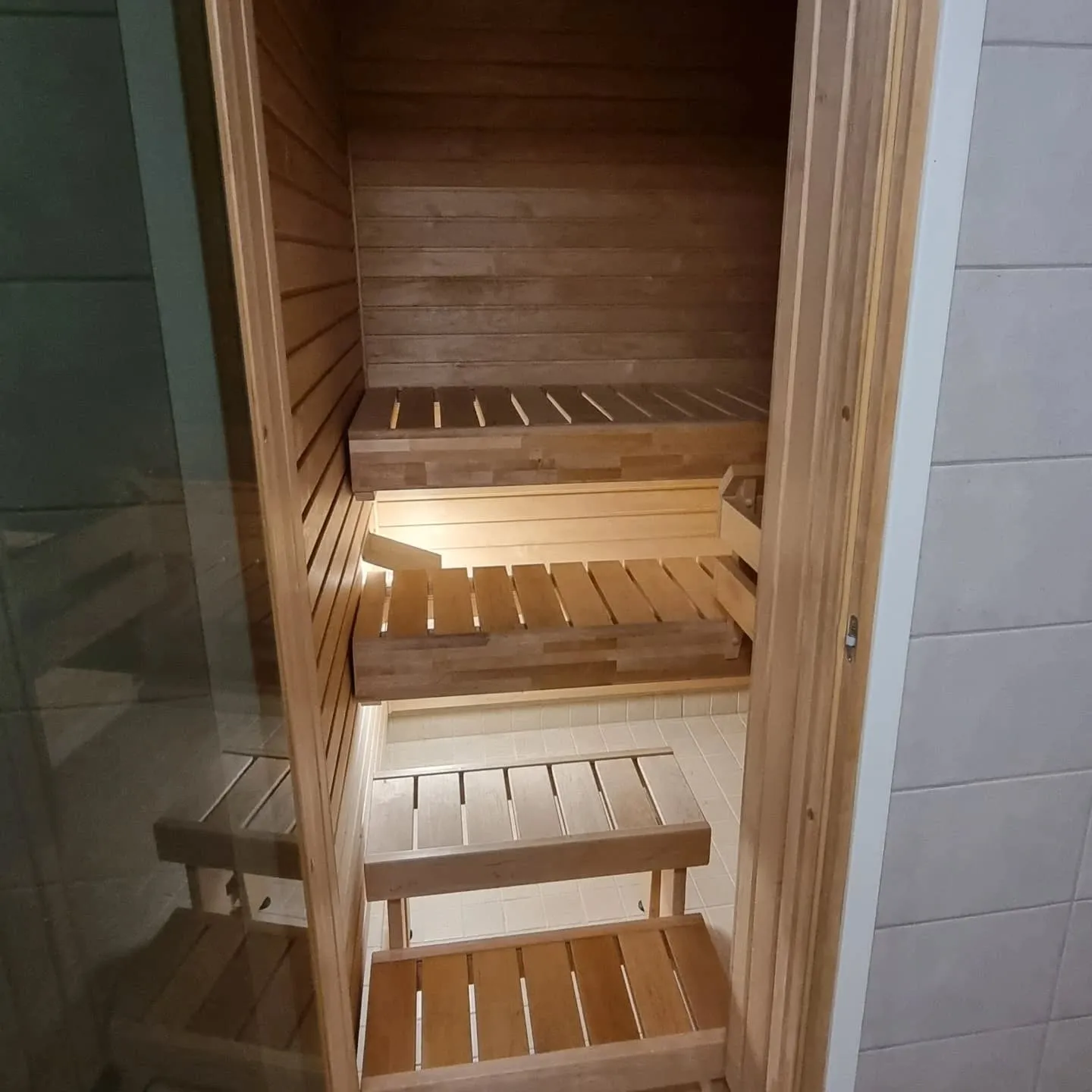 Kuva esittää modernin saunan sisätilaa, jossa on vaaleat puuseinät ja kolme tasoa puisia lauteita. Lämmin valaistus heijastuu lauteiden alta. Vasemmalla on lasinen ovi ja oikealla vaalea laattaseinä.