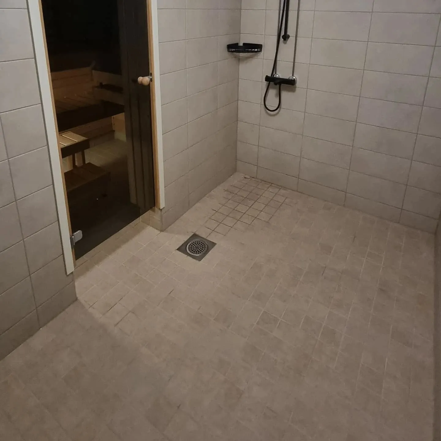 Vaaleanruskeilla laatoilla päällystetty suihkuhuone, jossa on saunan sisäänkäynti vasemmalla. Lattiassa on neliönmuotoinen lattiakaivo. Oikealla seinällä on musta suihku ja hylly. Saunan lasioven läpi näkyy puisia lauteita. Valaistus on hämärä.