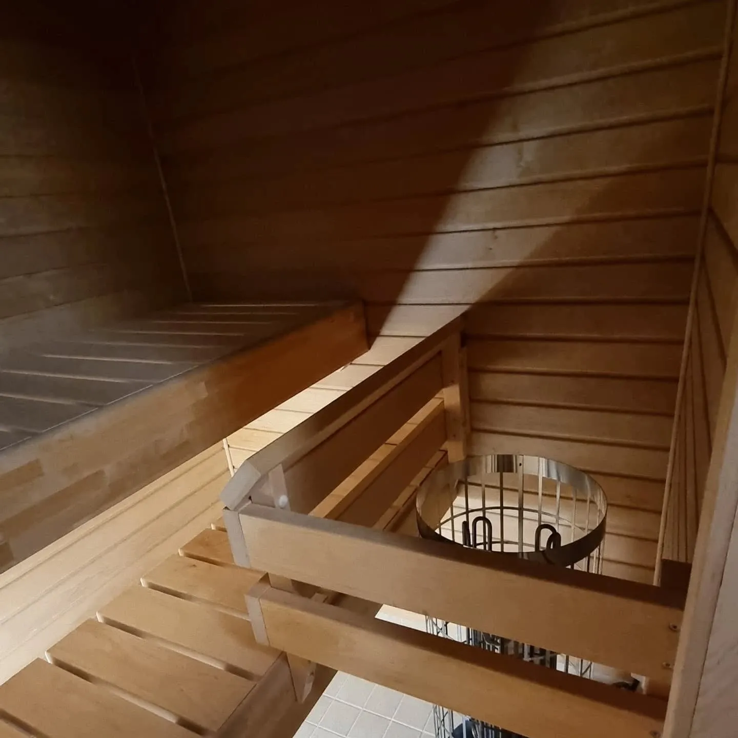 Saunan sisätila, jossa on vaaleat puuseinät ja kaksi puupenkkitasoa. Oikealla alhaalla on metalliverkolla suojattu sähkökiuas. Valo luo pitkän varjon takaseinälle ja ylemmälle penkille.