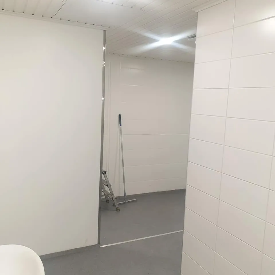 Sisätila, jossa on valkoiset seinät ja harmaa lattia. Vasemmalla on sileä valkoinen seinä, jonka reunassa on pystysuora metallilista. Oikealla on valkoinen kaakeloitu seinä. Takaseinä on valkoinen ja vaakasuuntaisesti paneeloitu. Katossa on valkoiset paneelit ja kirkas valo. Lattialla on valkoinen viiva. Vasemman seinän takana näkyy pienet tikkaat ja lattianpesuväline. Kuvan vasemmassa alakulmassa on osittain näkyvissä pyöreä valkoinen esine.