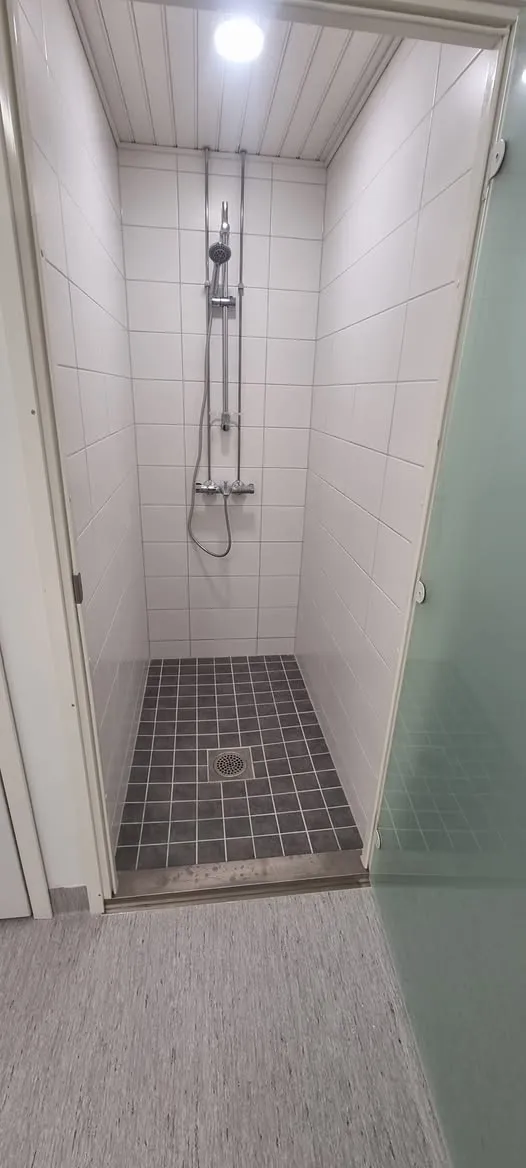 Suihkukaappi, jossa on valkoiset seinälaatat ja tummanharmaat neliönmuotoiset lattialaatat lattiakaivolla. Katossa on valkoiset paneelit ja kirkas pyöreä valaisin. Suihkussa on krominvärinen suihkupää ja termostaattihana. Suihkun edessä on vaaleanharmaa kuvioitu lattia ja oikealla himmeä lasiseinä.