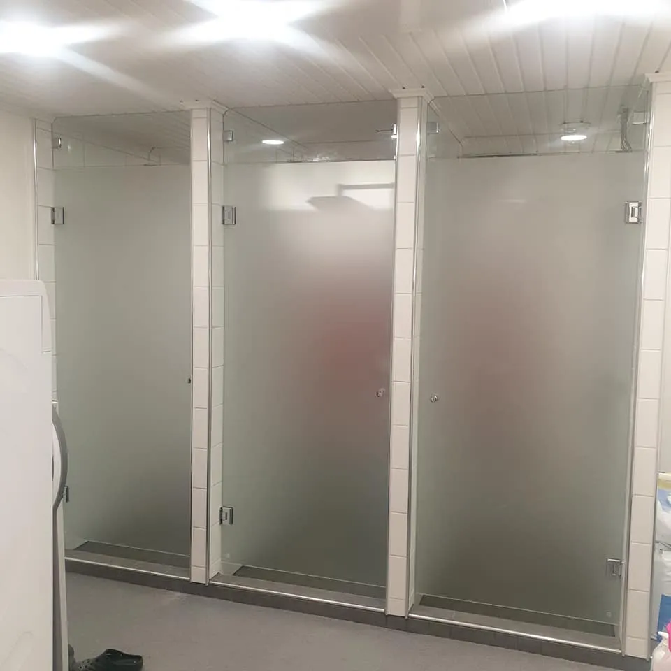Kolme vierekkäistä suihkukoppia, joissa on himmeät lasiovet ja valkoiset laattakehykset. Kopit seisovat harmaalla lattialla. Valkoinen paneelikatto upotettuine valoineen valaisee tilaa kirkkaasti. Vasemmalla näkyy osittain valkoinen kodinkone ja lattialla musta kenkä. Oikealla on joitakin siivoustarvikkeita.