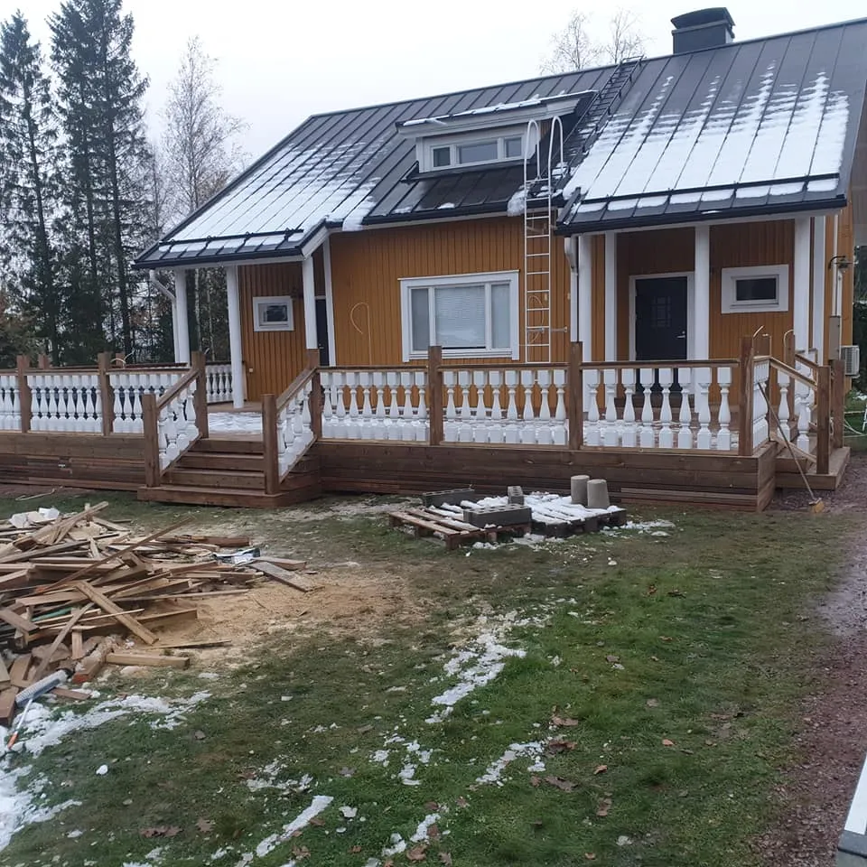 Pilvisenä päivänä otettu kuva keltaisesta puutalosta, jonka tummalla peltikatolla on lumipeite. Talon edessä on laaja puinen terassi valkoisilla kaiteilla ja portailla. Maassa on vihreää ruohoa, lumiläiskiä ja puunpalasia. Katon harjalla on valkoiset tikkaat. Taustalla näkyy havupuita ja lehtipuita.