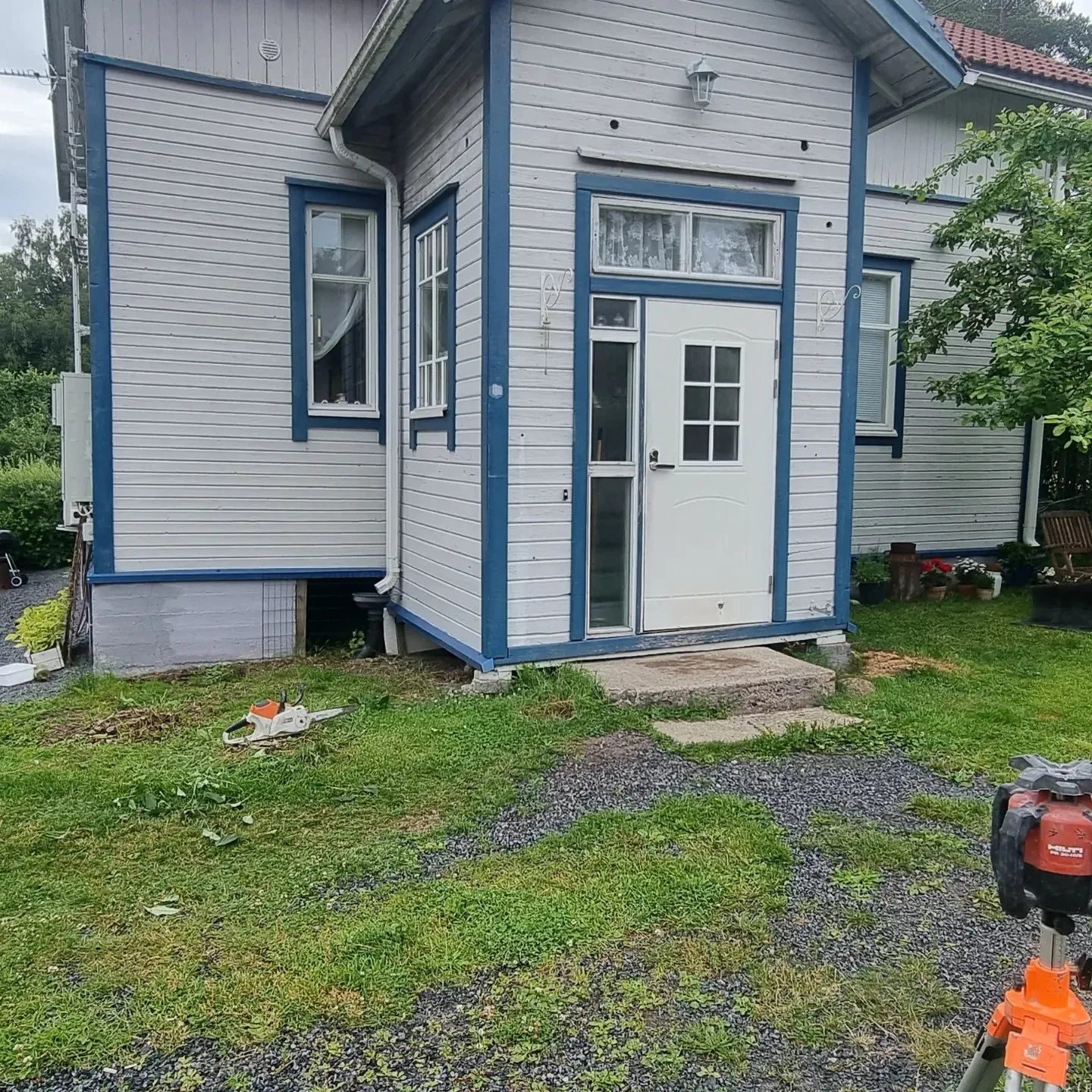 Vaaleanharmaa puutalo sinisillä ikkunan- ja ovenkarmeilla sekä kulmalistoilla. Talon edessä on nurmikkoa ja tummanharmaata sorapolkua. Nurmikolla makaa oranssi-valkoinen moottorisaha. Oikealla etualalla on punainen laseretäisyysmittari oranssilla jalustalla. Talon sisäänkäynnin edessä on betoniporras. Kuva on otettu pilvisenä päivänä, valaistus on tasainen.