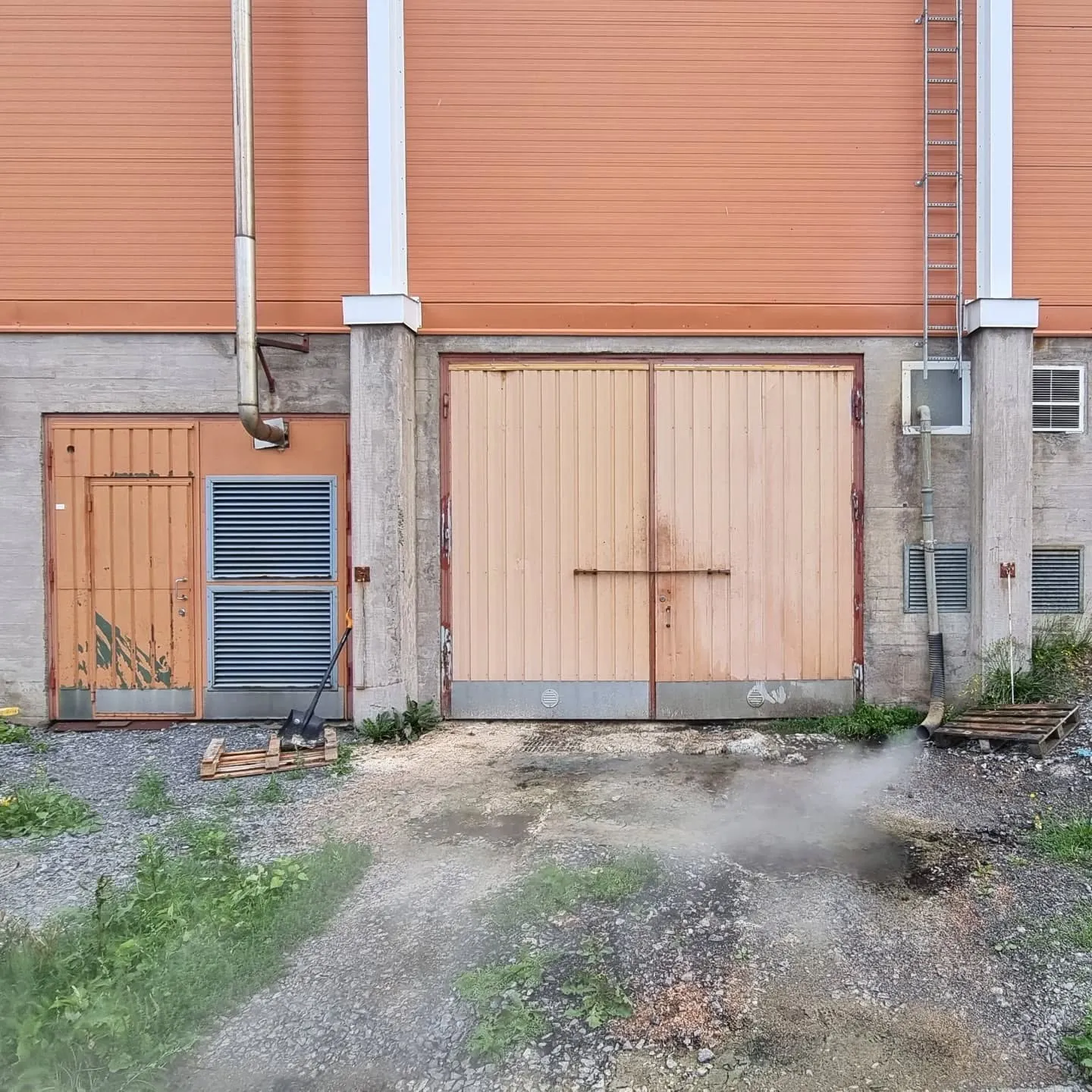 Teollisuusrakennuksen ulkokuva, jossa oranssi aaltopeltiseinä ja harmaa betonijalusta. Vasemmalla on oranssi ovi sinisen ilmanvaihtoritilän vieressä ja pitkä metallipiippu. Keskellä on suuret vaaleanoranssit pariovet. Oikealla seinällä on metalliset tikkaat ja pienempiä ilmanvaihtoritilöitä. Maassa on soraa, ruohoa ja kaksi puulavaa. Etualalla maasta nousee savua tai höyryä.