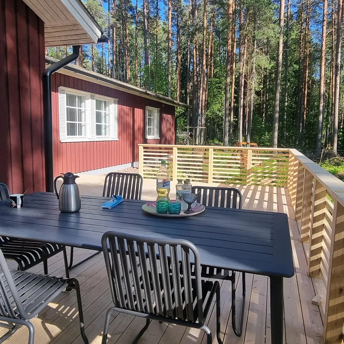Aurinkoisena päivänä punaisen puutalon vieressä on puinen terassi. Terassilla on tummanharmaa suorakulmainen pöytä ja useita samanlaisia tuoleja. Pöydällä on hopeinen termospullo, sininen esine, tarjotin kahdella turkoosilla lasikynttilänjalalla ja harmaalla lasimaljalla sekä kaksi pulloa. Talon seinässä on valkoiset ikkunat ja musta syöksytorvi. Terassia ympäröi vaalea puukaide, ja taustalla näkyy tiheä mäntymetsä sinistä taivasta vasten. Auringonvalo luo varjoja terassin lattialle.
