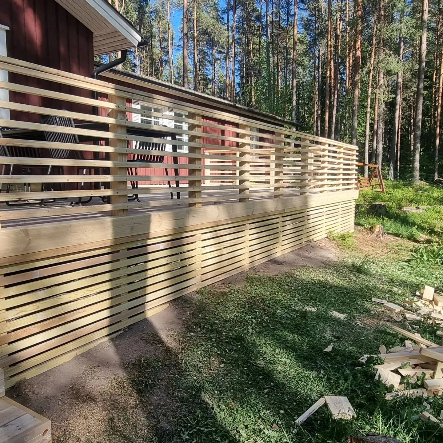 Aurinkoisena päivänä otettu kuva punaisesta talosta, jonka edessä on uusi, vaaleasta puusta rakennettu terassi. Terassin kaide ja alusrakenne on tehty vaakasuorista laudoista. Taustalla näkyy tiheä mäntymetsä. Maassa on vihreää nurmikkoa ja rakennustyöstä jääneitä puunpalasia. Terassilla näkyy tummia ulkokalusteita.