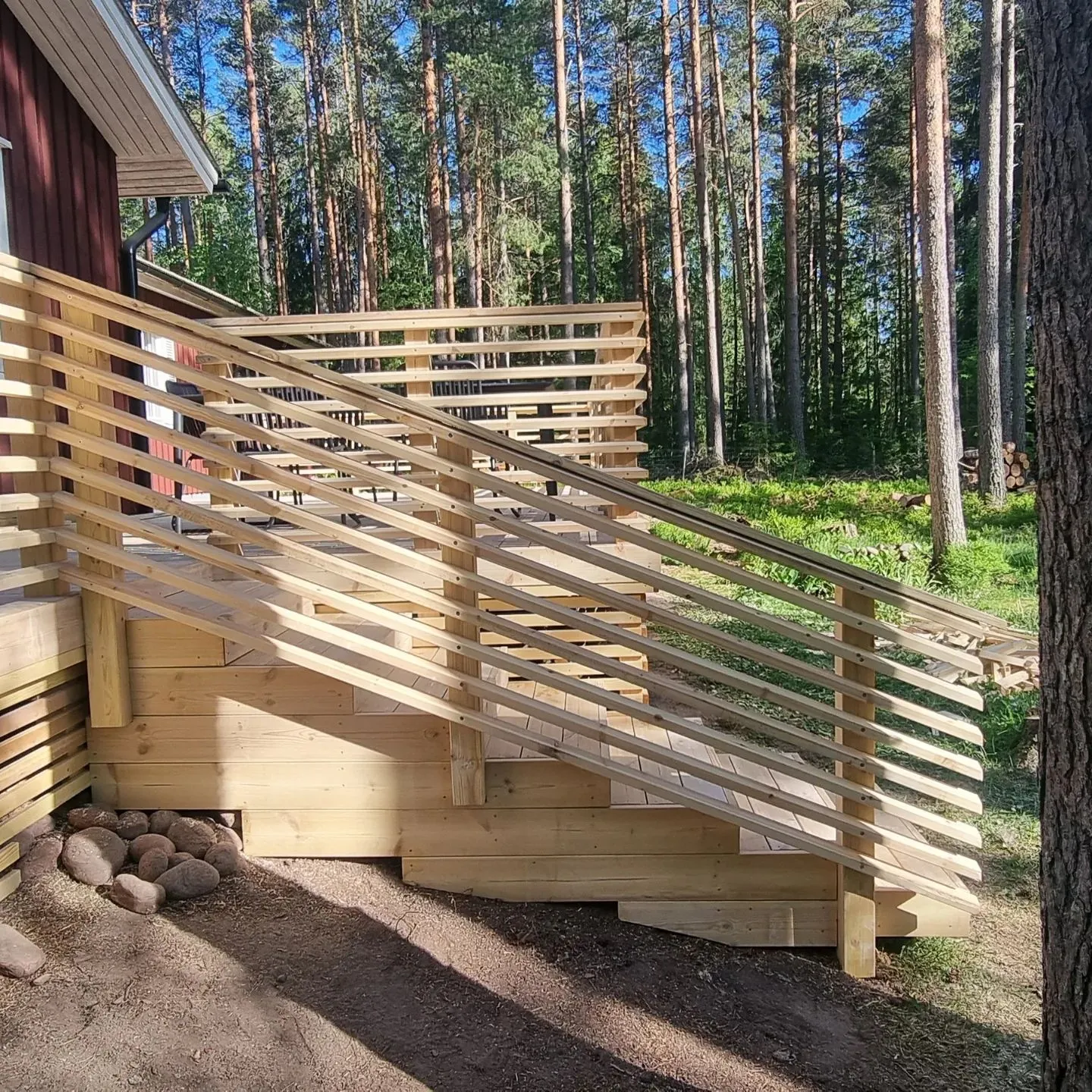 Uusi puinen terassi ja portaat johtavat ylös punaiselle talolle. Terassissa ja portaissa on vaaleat vaakasuorat puukaiteet. Taustalla on tiheä mäntymetsä ja kirkas sininen taivas. Maassa on kiviä ja pitkiä varjoja aurinkoisena päivänä.