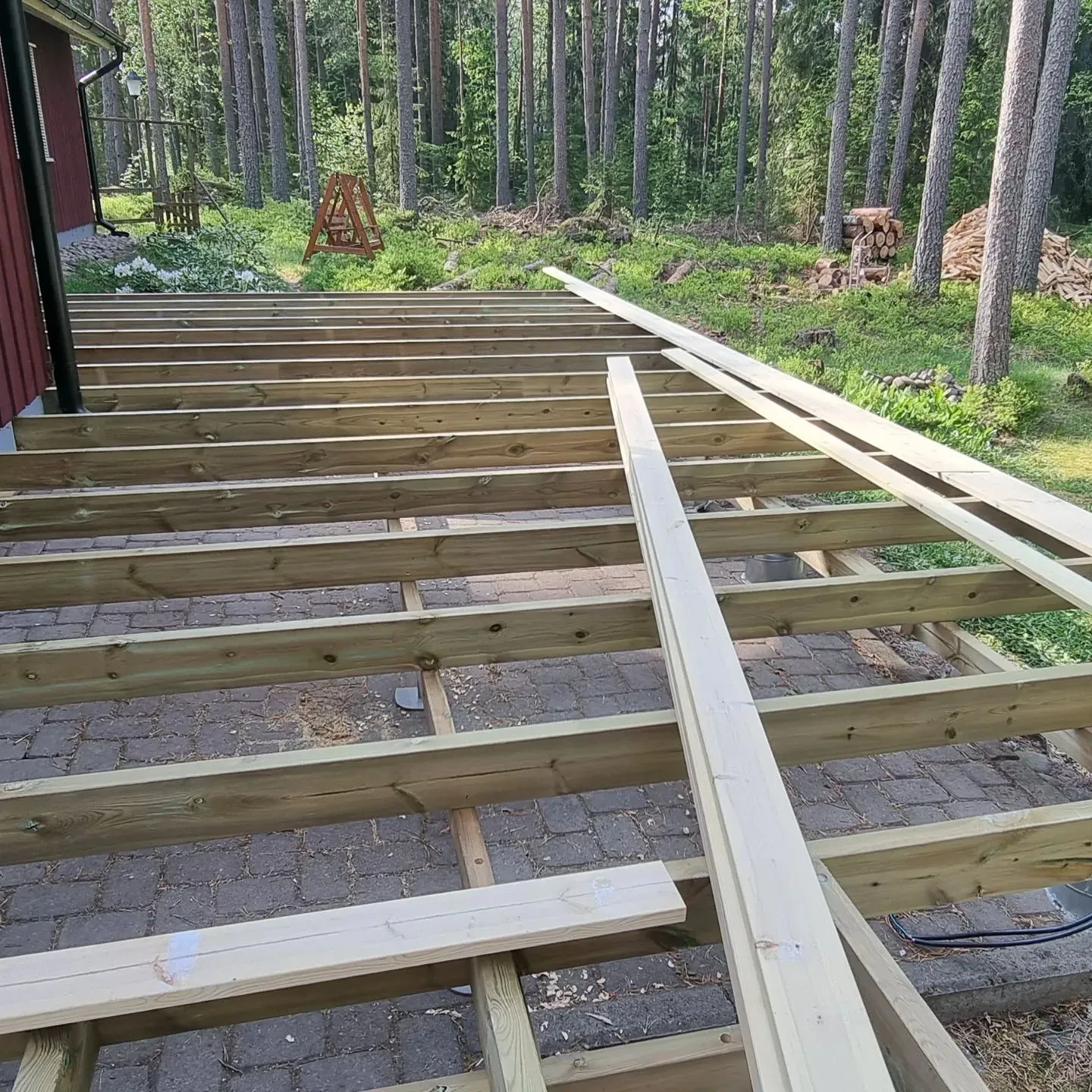 Rakenteilla oleva puinen terassin runko punaisen talon vieressä. Runko on tehty vihreästä painekyllästetystä puusta, ja vaaleita terassilautoja on aseteltu sen päälle. Taustalla näkyy tiheä mäntymetsä, jossa on polttopuukasoja ja puinen A-teline. Terassin alla on kivilaatoitus. Kuva on otettu aurinkoisena päivänä.