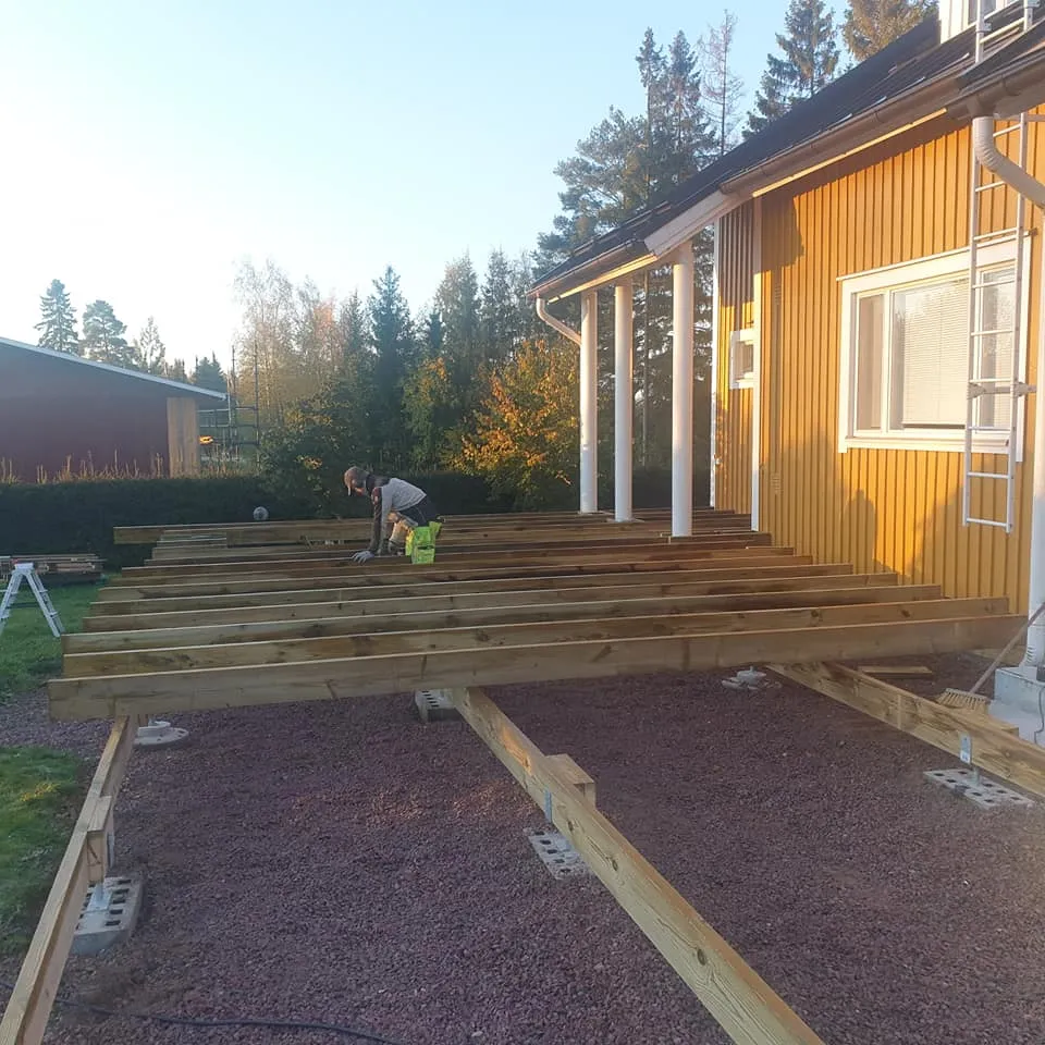 Henkilö rakentaa puista terassin runkoa keltaisen talon viereen. Terassin puupalkit on asetettu punertavalle sorapedille, ja ne lepäävät betoniharkkojen ja säädettävien jalkojen päällä. Keltaisessa talossa on valkoiset pylväät ja ikkuna. Taustalla näkyy puita syksyisissä väreissä kirkkaassa auringonvalossa.