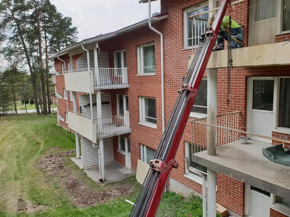 Punainen tiilitalo, jossa on useita valkoisia parvekkeita. Oikealla ylhäällä työntekijä vihreässä paidassa ja tummissa housuissa työskentelee parvekkeella. Punainen nosturi ulottuu parvekkeen tasolle. Taustalla näkyy vihreää nurmikkoa, muokattua maata ja korkeita mäntyjä pilvisessä säässä.