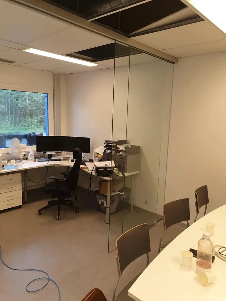 Valoisa toimistotila, jossa vasemmalla on työpöytä kahdella näytöllä ja mustalla työtuolilla. Ikkunasta näkyy puita. Lasiseinä jakaa tilan, ja oikealla on valkoinen kokouspöytä tuoleineen, vesipullo ja kuppeja. Katossa on alaslaskettu rakenne, josta puuttuu paneeleja, ja loisteputkivalaisimia. Lattialla on harmaa matto.