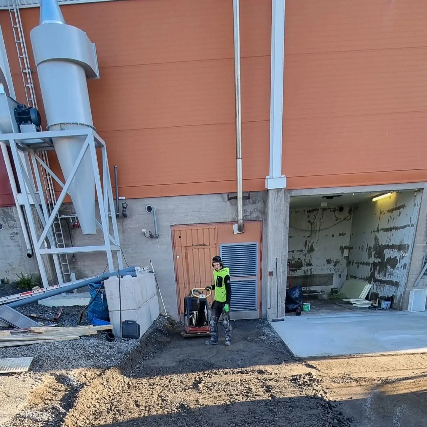 Henkilö kirkkaanvihreässä takissa ja kuulokkeissa tiivistää maata täryttimellä rakennustyömaalla. Taustalla on suuri vaaleanharmaa teollisuussäiliö, oranssinruskea rakennuksen seinä ja avoin betoninen sisäänkäynti. Maa on soraa ja multaa, ja oikealla on uusi betonilaatta. Aurinkoinen päivä.