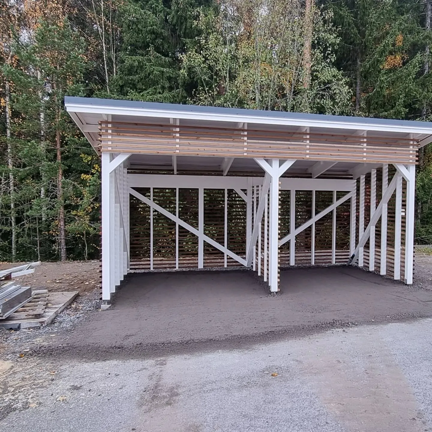 Uusi, valkoinen puukatos harmaalla katolla ja vaaleilla puurimoilla. Katoksessa on kaksi ajoneuvopaikkaa ja sorapohja. Taustalla on syksyinen metsä ja edessä hiekkatie. Vasemmalla näkyy rakennustarvikkeita.
