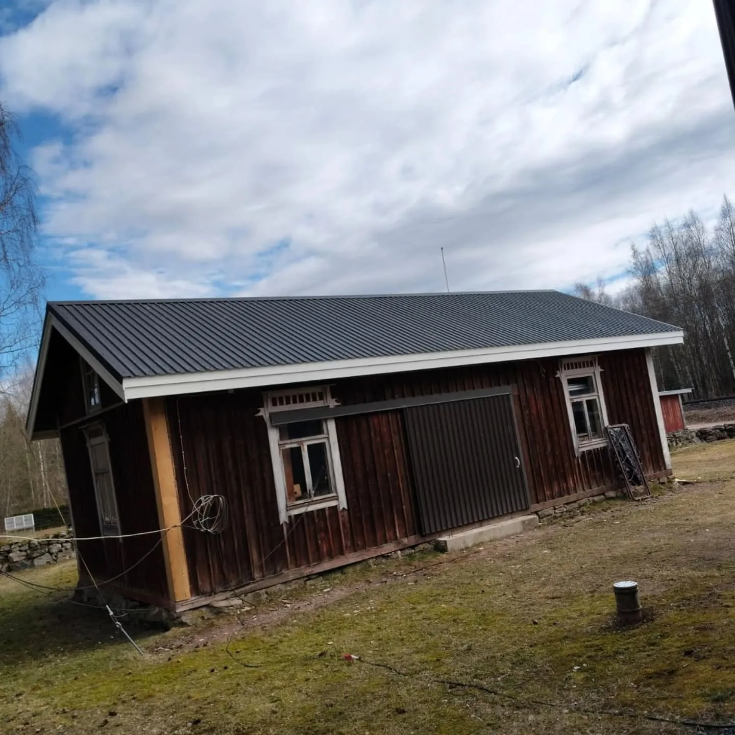 Vanha tummanpunainen puurakennus tummanharmaalla peltikatolla. Rakennuksessa on valkoiset ikkunanpielet ja räystäät, sekä tummanruskea liukuovi. Vasemmalla puolella on vaaleampi pystylauta ja maahan kiinnitettyjä johtoja. Maaperä on kuivaa ruohoa, jossa on vihreitä laikkuja. Taustalla on paljaita puita ja kirkas sininen taivas valkoisine pilvineen. Kuva on otettu hieman vinossa kulmassa.